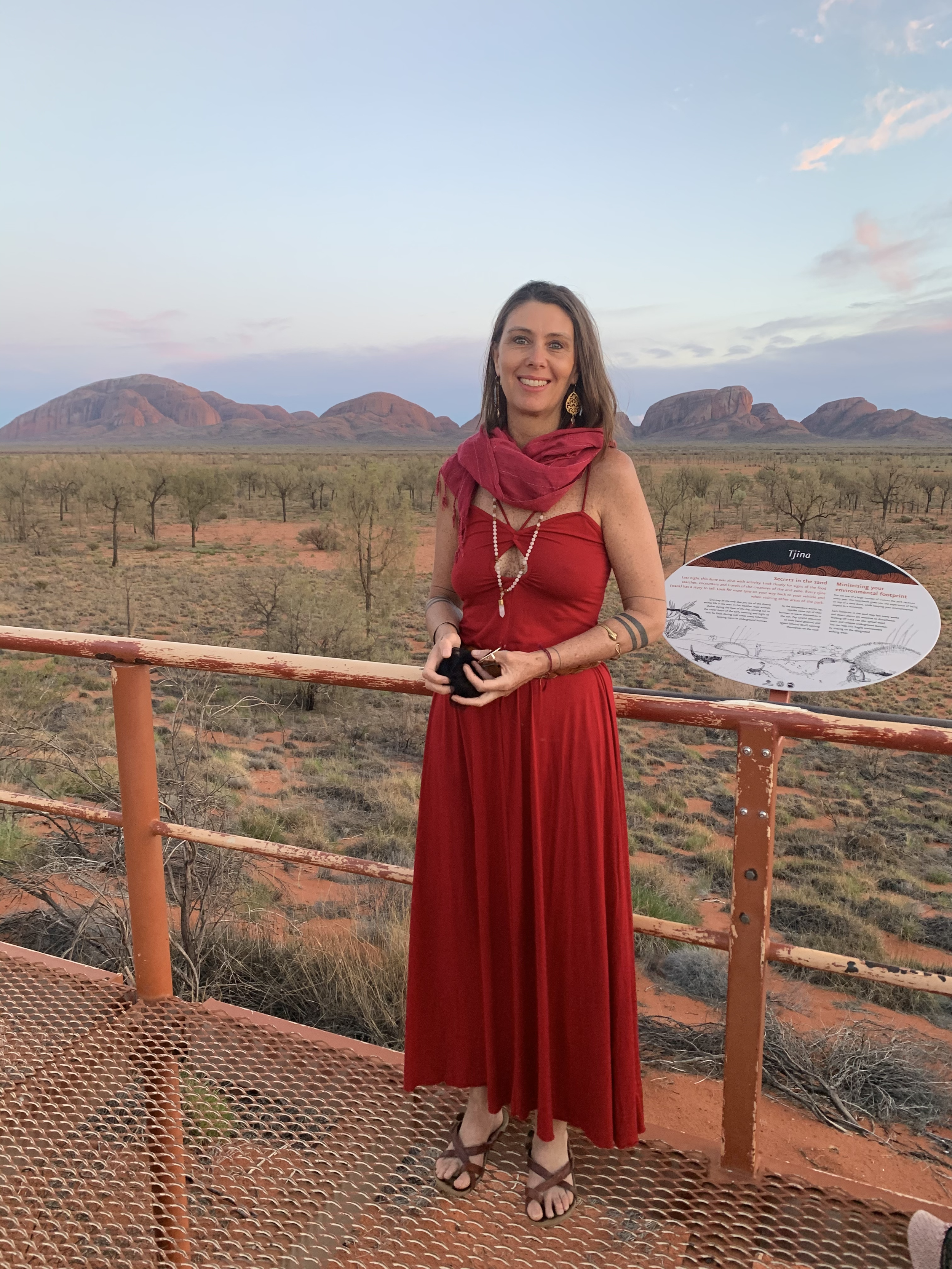 A woman in a red dress and scarf standing on a viewing platform in a desert landscape with mountains in the background.