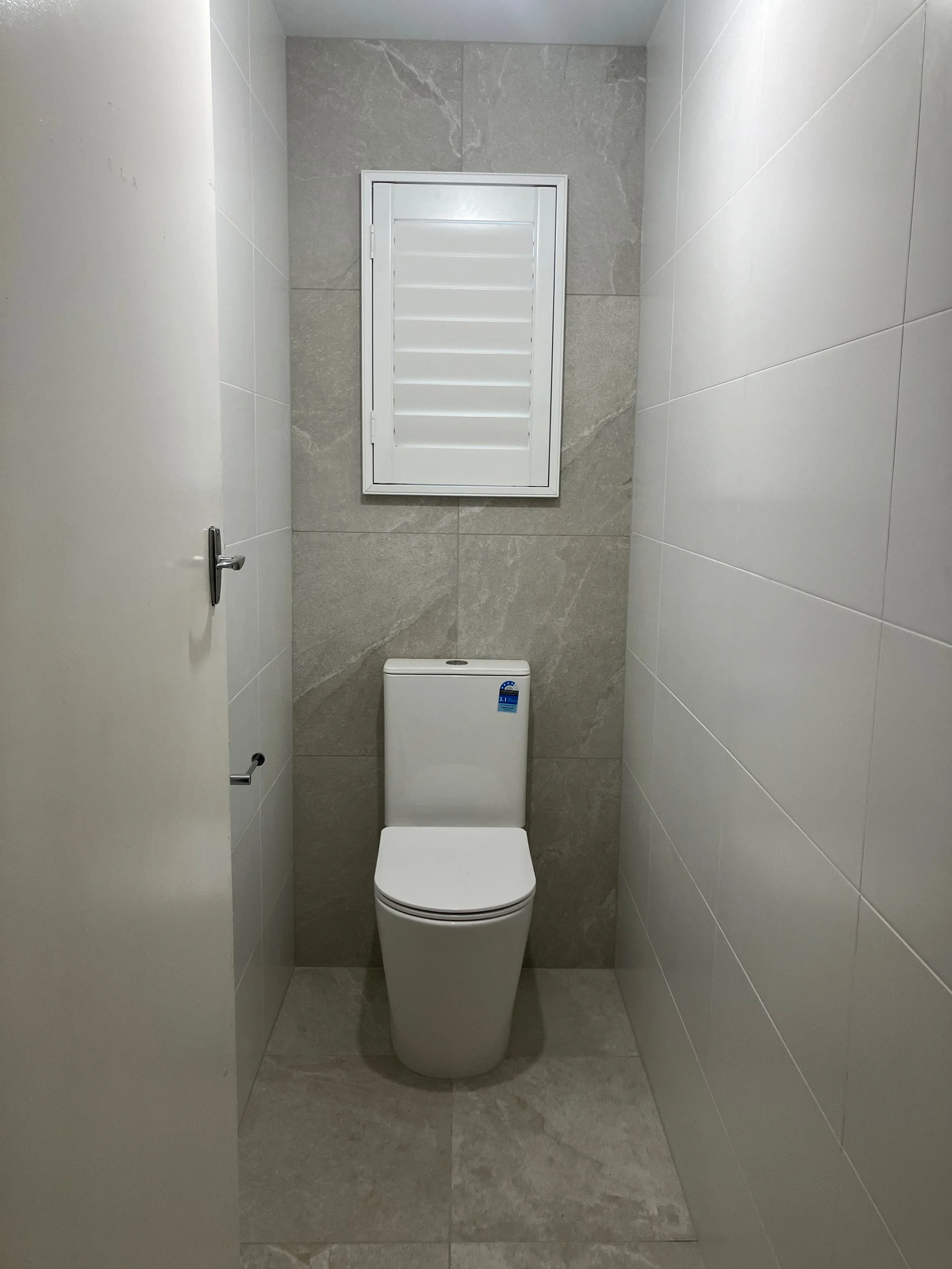 A small, modern bathroom with beige and gray tiled walls and floor, containing a white toilet and a small window with horizontal blinds.