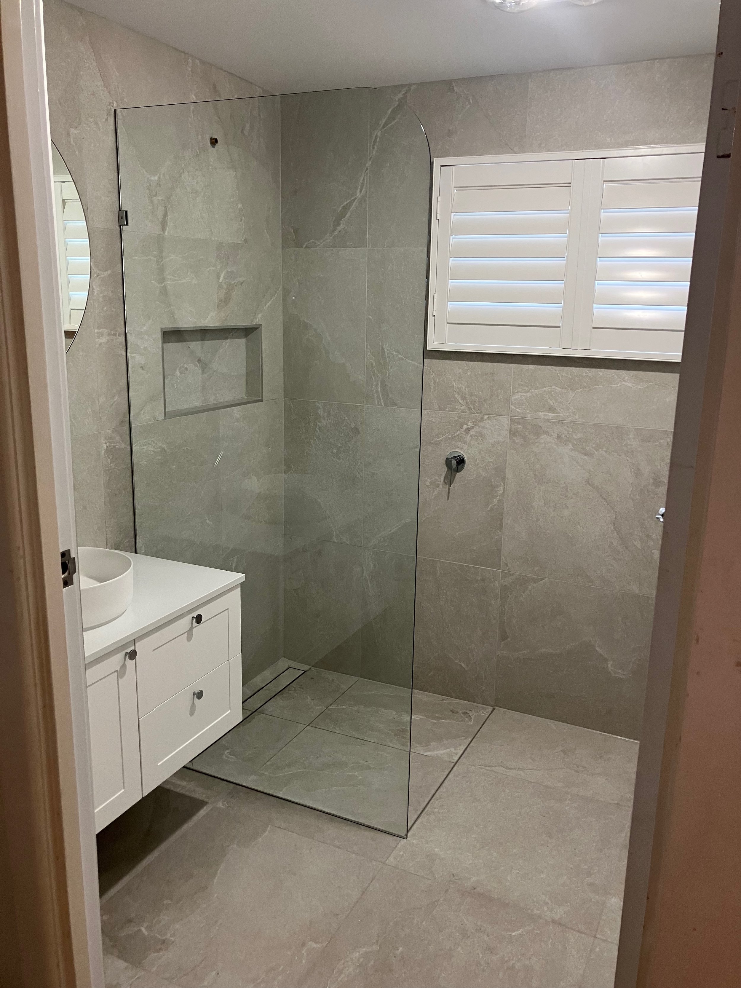 A modern bathroom with beige marble tiles on the floor and walls, a white vanity with a bowl sink, a glass shower enclosure with a built-in niche, a wall-mounted shower fixture, and a window with white shutters.