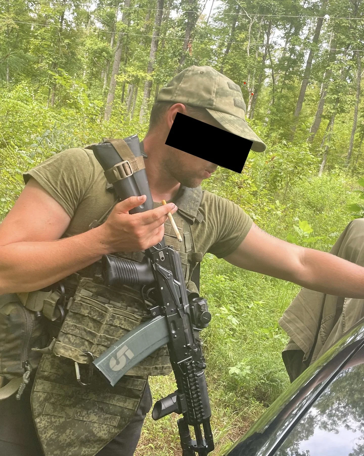 A man in military gear, including a camouflage shirt and cap, is outdoors in a wooded area. He is holding a handgun and a cigarette, with a rifle slung over his shoulder, next to a black vehicle.
