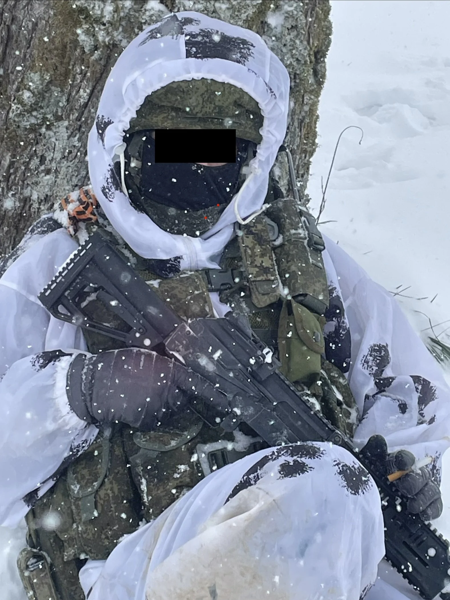 A soldier dressed in camouflage gear with a white weather-resistant cover, holding a black rifle, sitting against a tree in a snowy environment with snow falling.