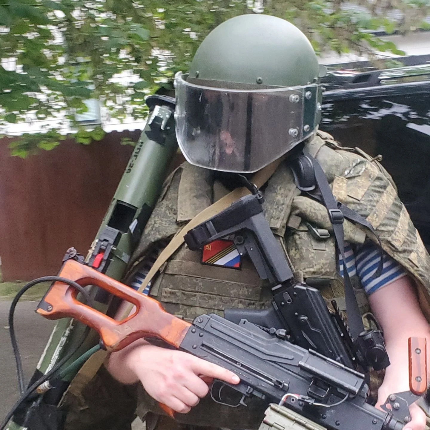 A person in military gear wearing a helmet with face shield, camo vest, and striped shirt, holding a firearm and a rocket launcher.