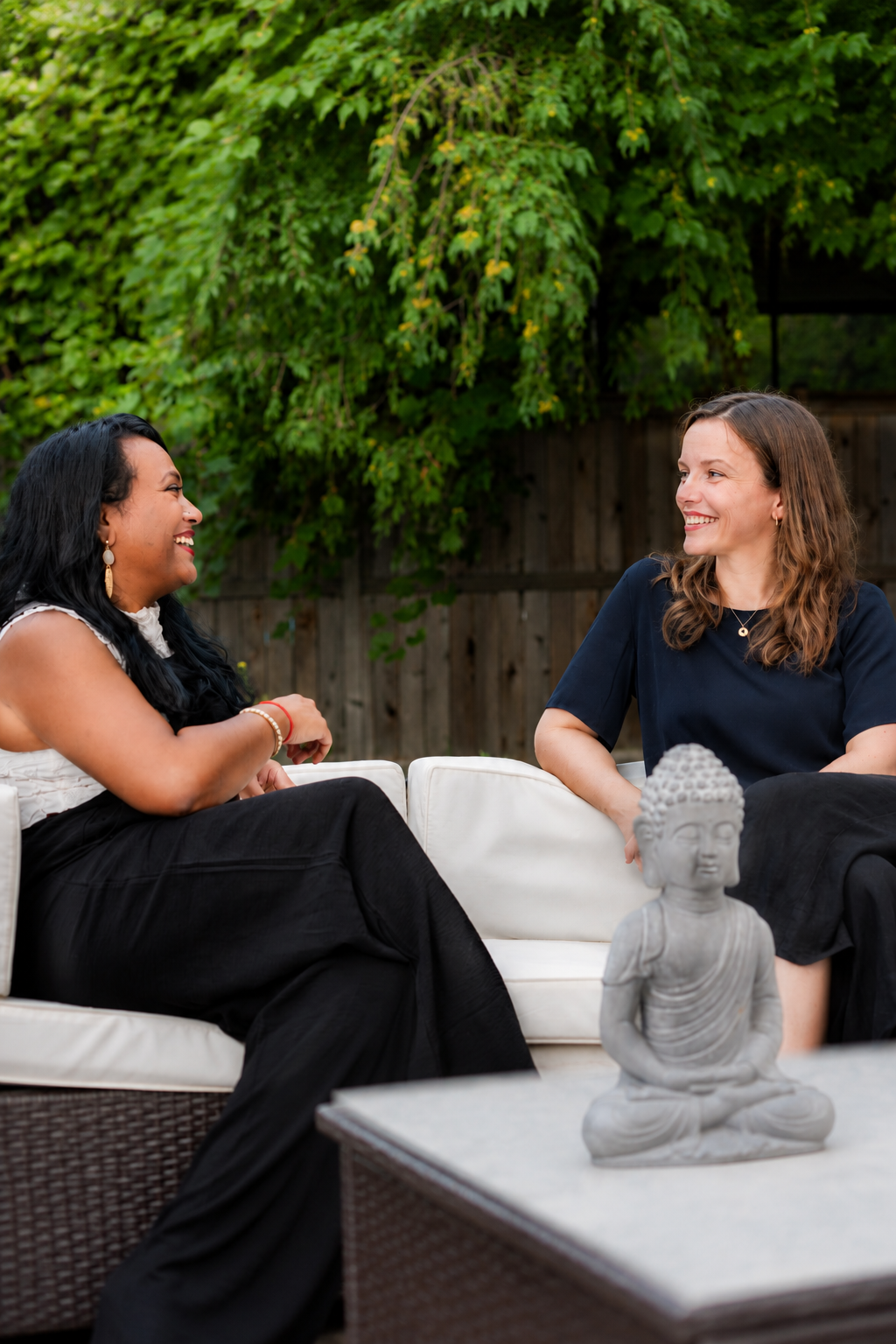 Two women sitting on a white outdoor sofa, smiling and talking, with a Buddha statue on the table in front of them and green foliage in the background.