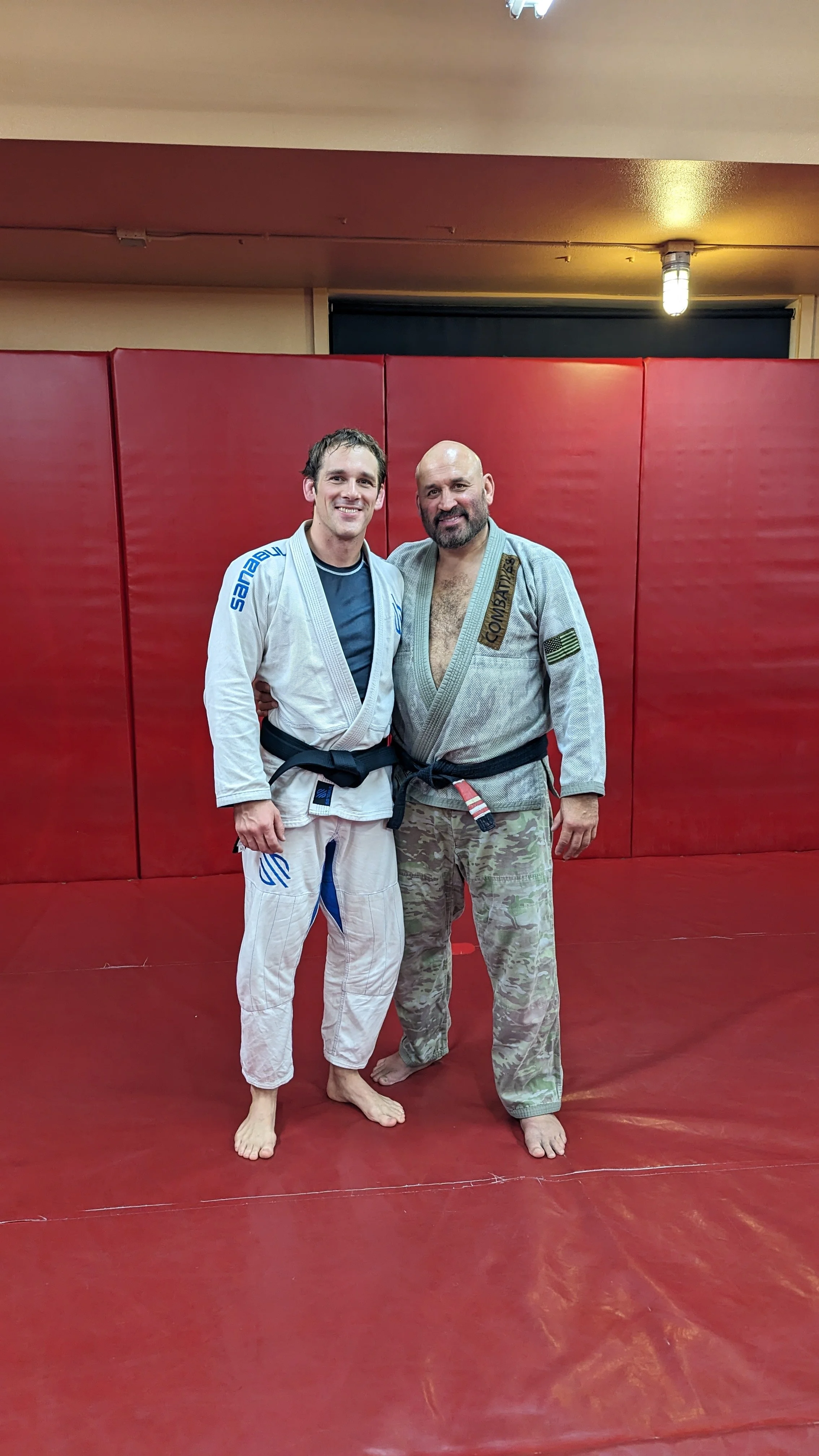 Two men in Brazilian Jiu-Jitsu gis standing together on a red mat, smiling at the camera. One man is wearing a white gi, the other a gray gi with camouflage pants.