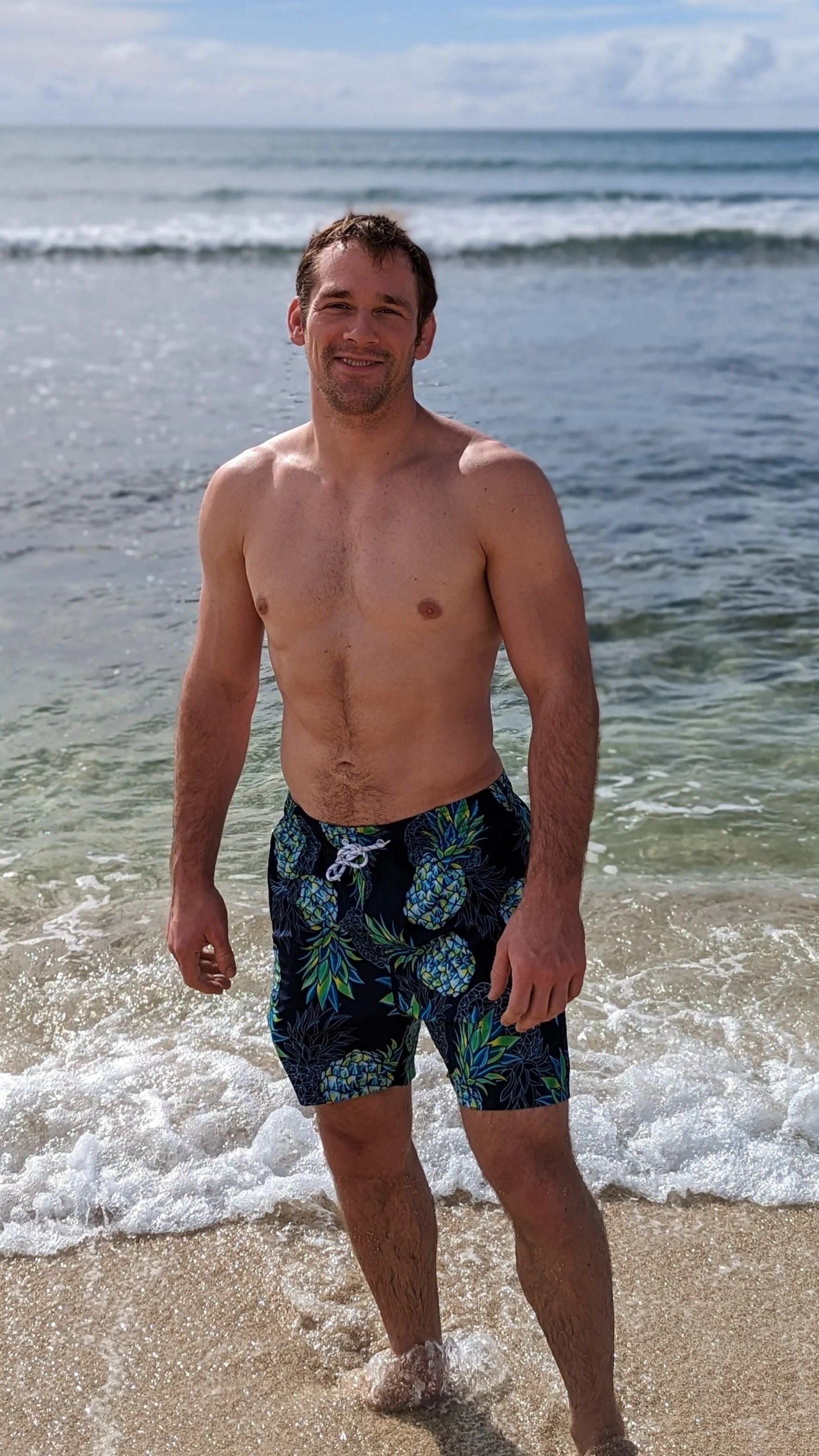 A man standing in the ocean with a smile, wearing blue and green pineapple-patterned swim trunks, with waves and a cloudy sky in the background.