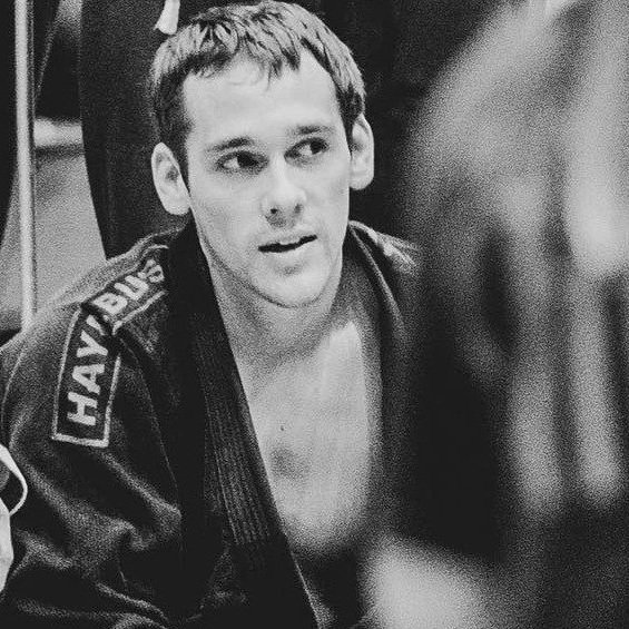 A young man with short hair and a serious expression, wearing a jiu-jitsu gi, sitting in a training environment.