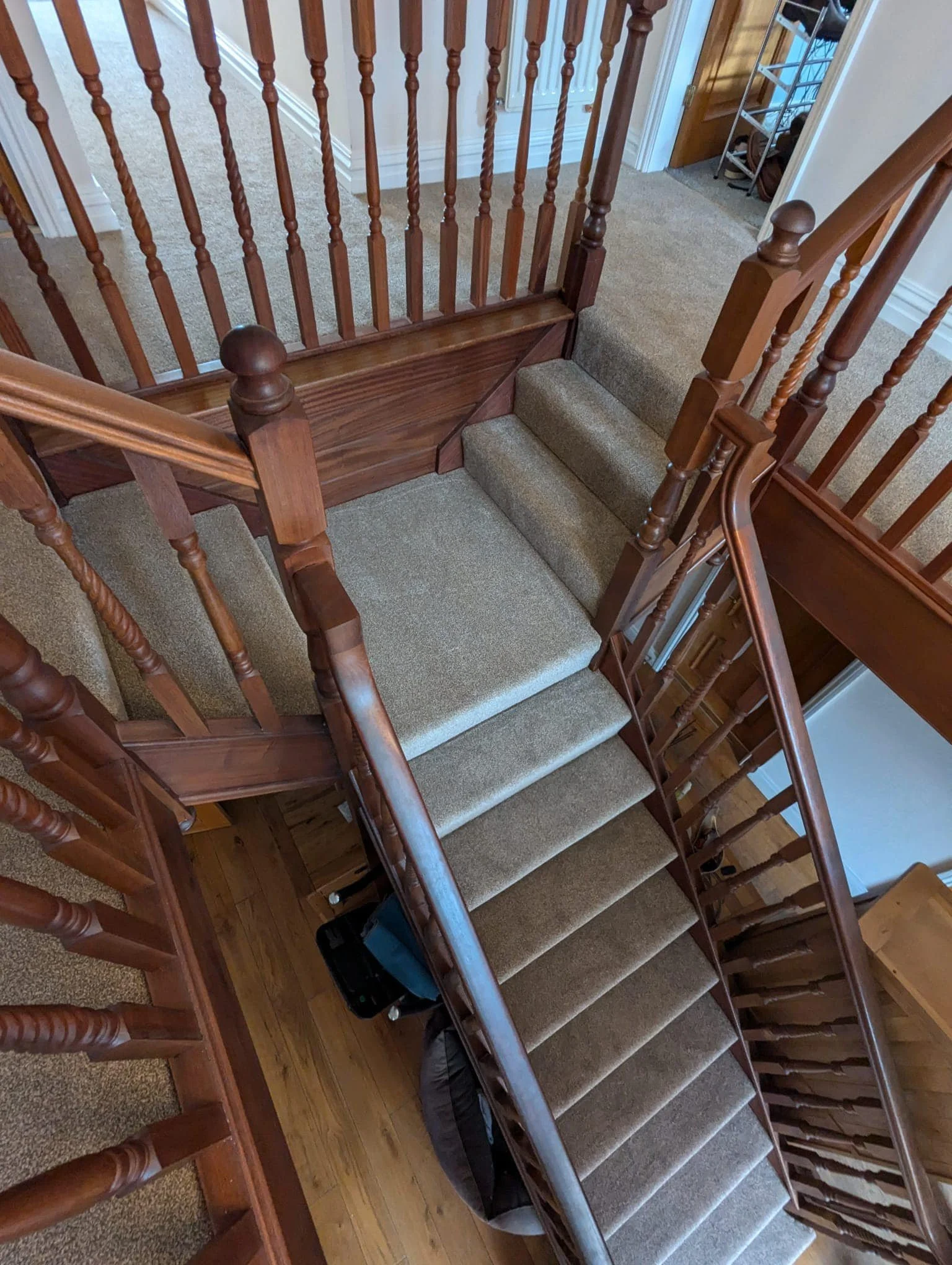 View of a staircase with carpeted steps, wooden handrails, and spindles, leading down to a lower floor with wooden flooring. There's a small black bag at the bottom of the stairs.