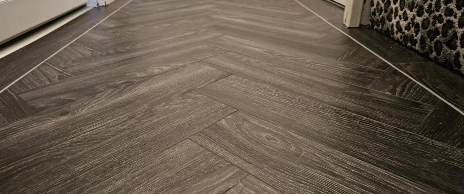 Close-up view of a wood-look herringbone-patterned vinyl or laminate flooring in a room corner.