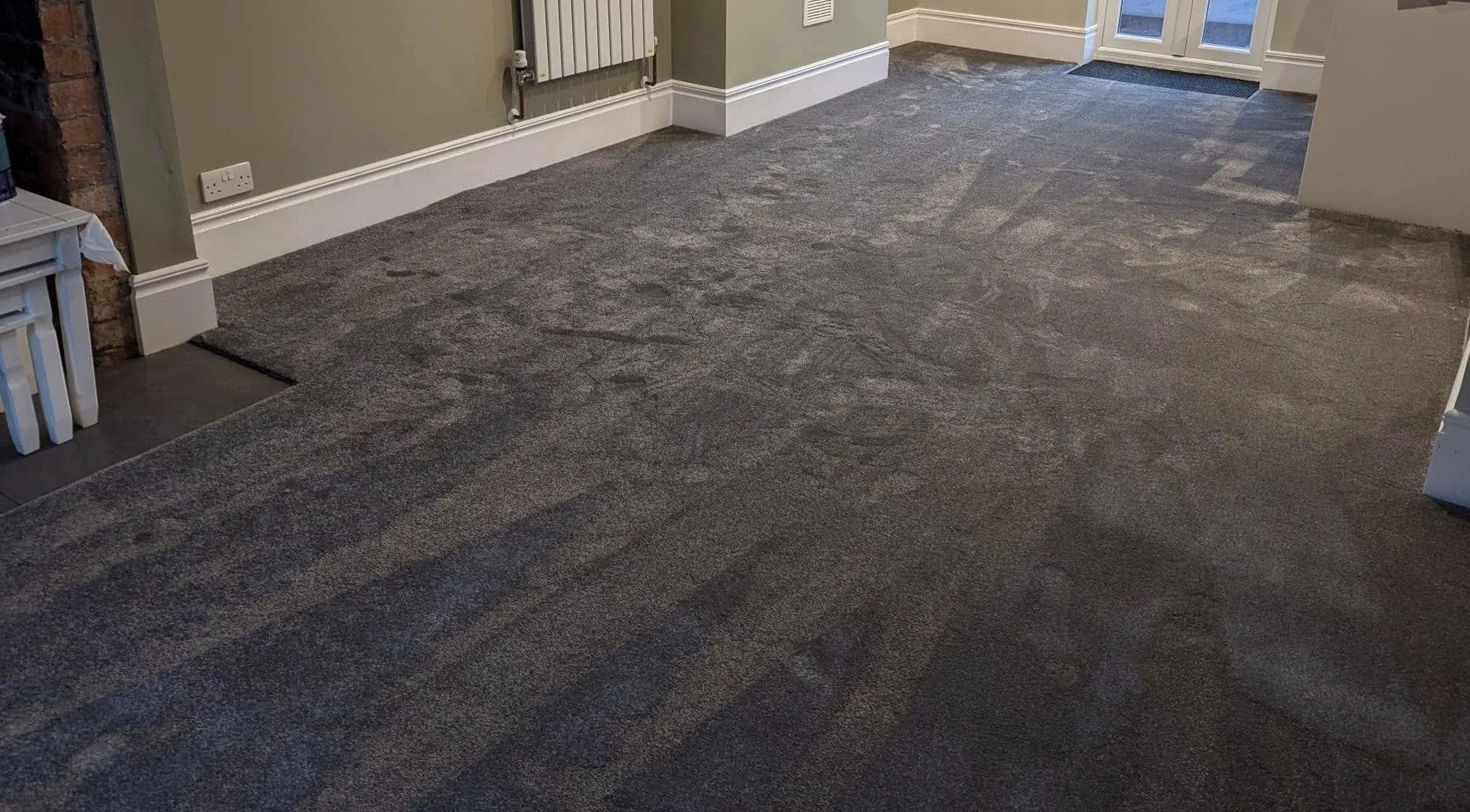 Room with gray carpet flooring, white baseboards, a green wall, a radiator, and a glass door at the far end.