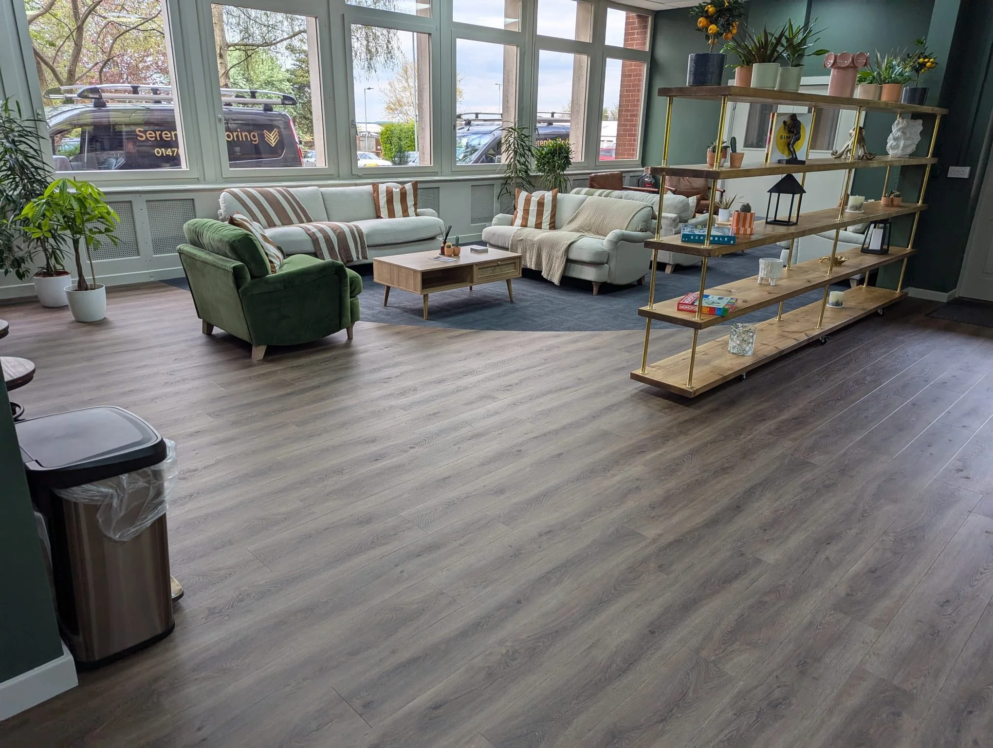 Modern living room with large windows, white and green sofas, a wooden coffee table, and a gold-framed bookshelf with decorative items, plants, and books.