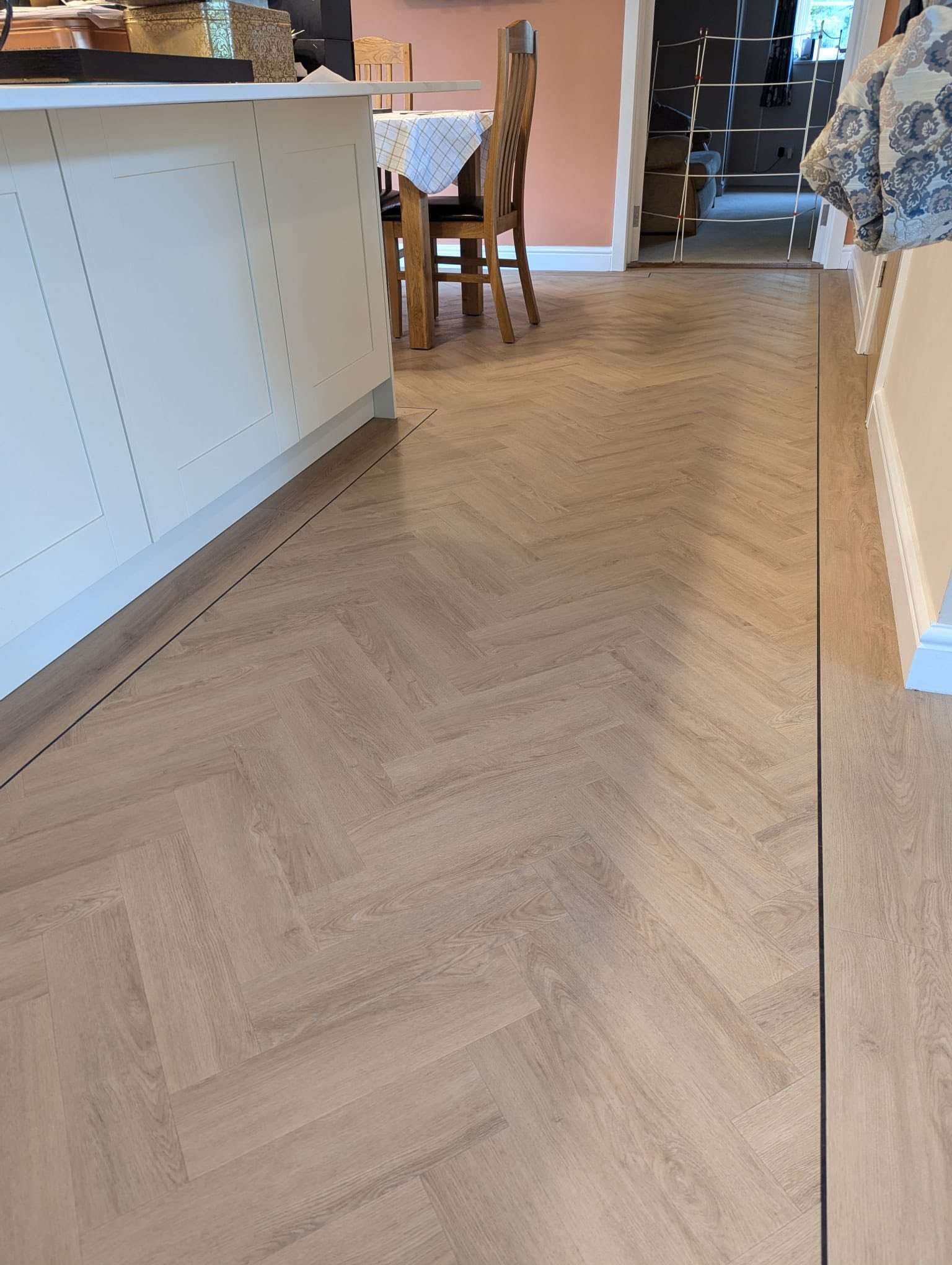 Long wooden flooring with a herringbone pattern in a kitchen and dining area, with white cabinets, wooden chairs, and a table with a checkered tablecloth.