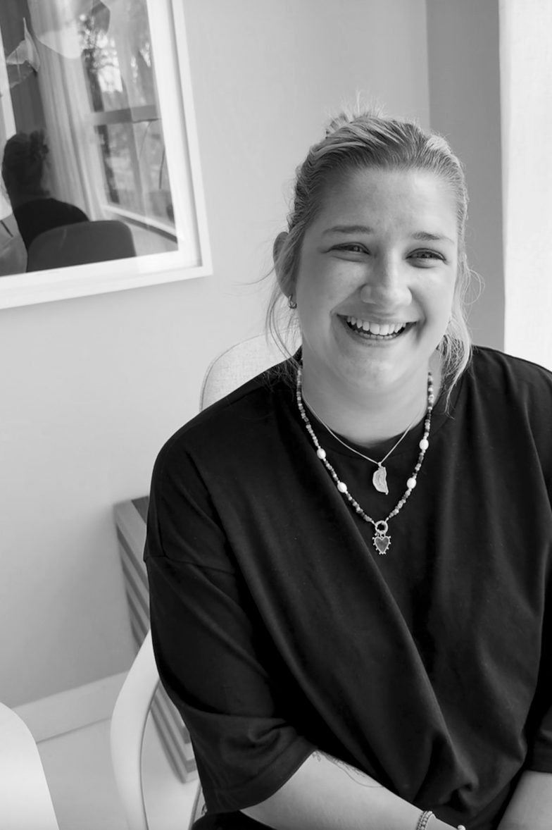 A smiling young woman with light hair in a ponytail, wearing a dark shirt and layered necklaces, sitting indoors near a window with a reflection of a person.
