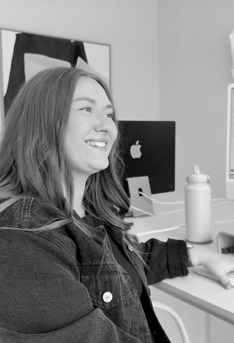 A young woman with long hair smiling while working on a laptop at a desk in a modern office.