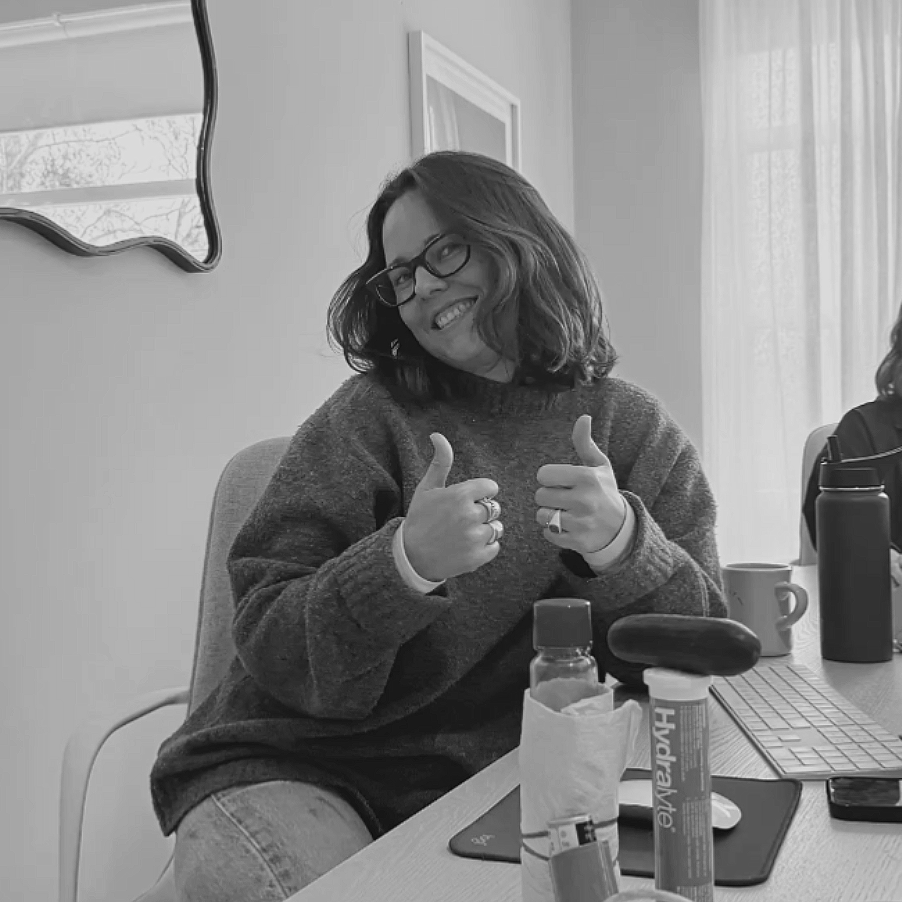 A woman with shoulder-length hair, glasses, and a sweater, smiling and giving two thumbs up while sitting at a table in a well-lit room.