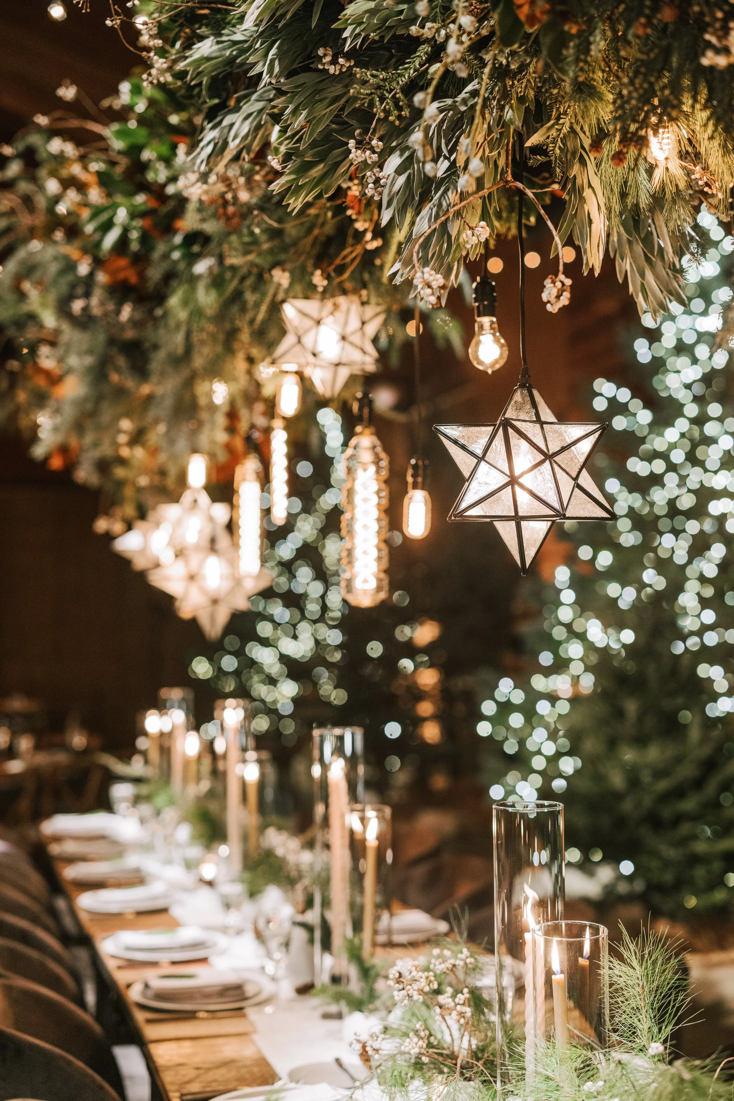 A festive dining table decorated with candles, greenery, and white flowers, with hanging star-shaped and lantern-style lights overhead. In the background, lights and a Christmas tree are visible.