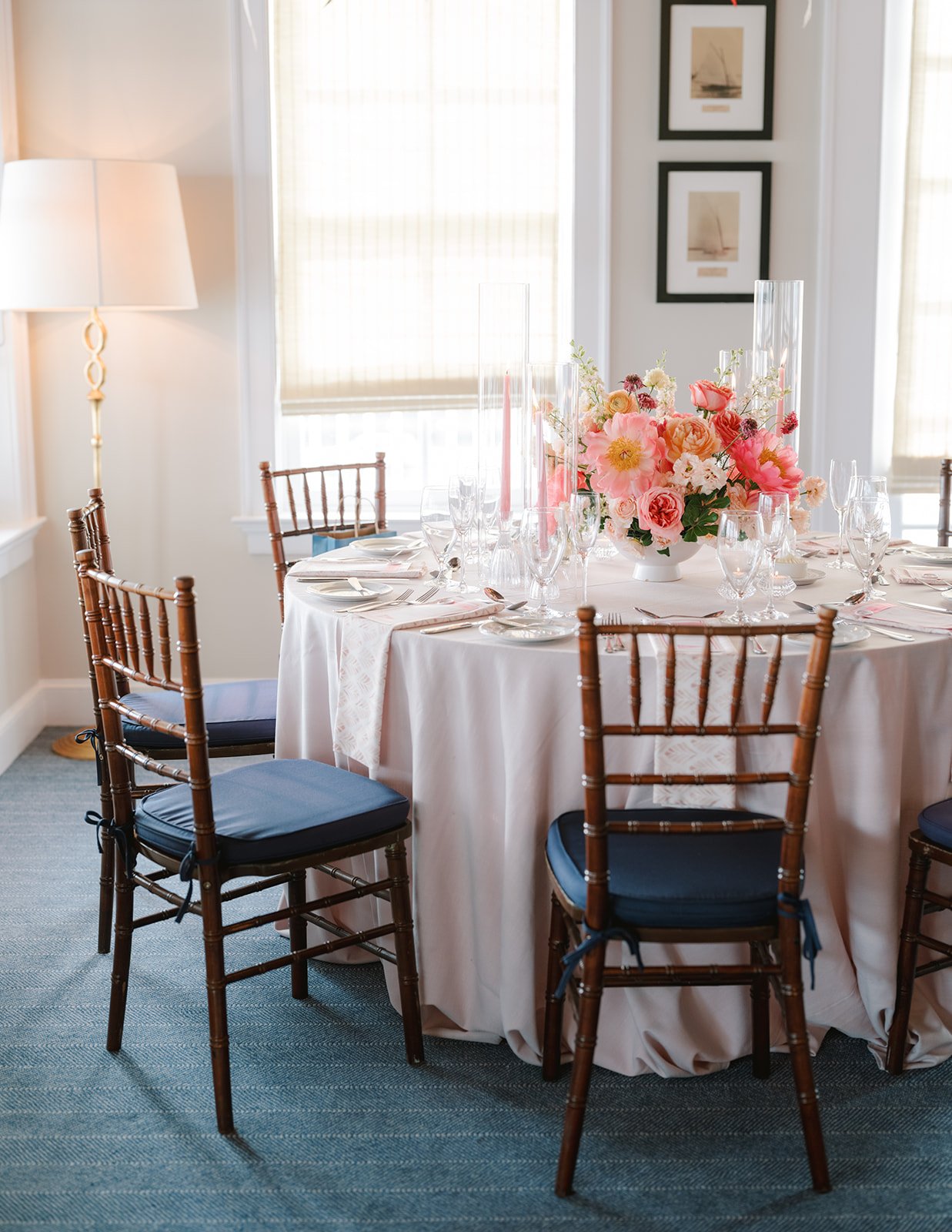 A round banquet table set for a formal event with a white tablecloth, pink floral centerpiece, and glassware. Wooden chairs with blue cushions surround the table, with artwork hanging on the wall and a lamp beside the window in the background.