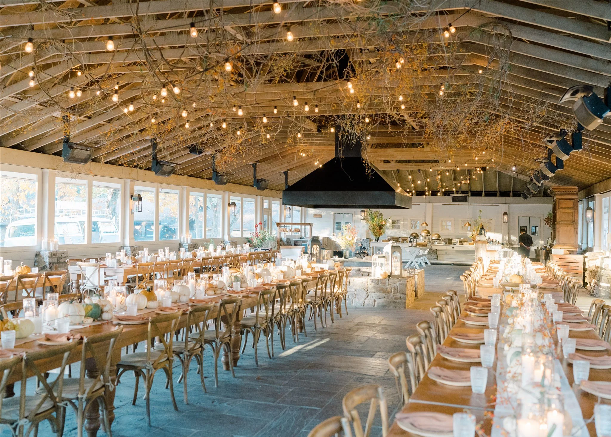 Interior of a decorated banquet hall with long wooden tables, candles, pumpkins, and fall-themed centerpieces, set for a gathering or celebration, with string lights hanging from the ceiling and large windows letting in natural light.