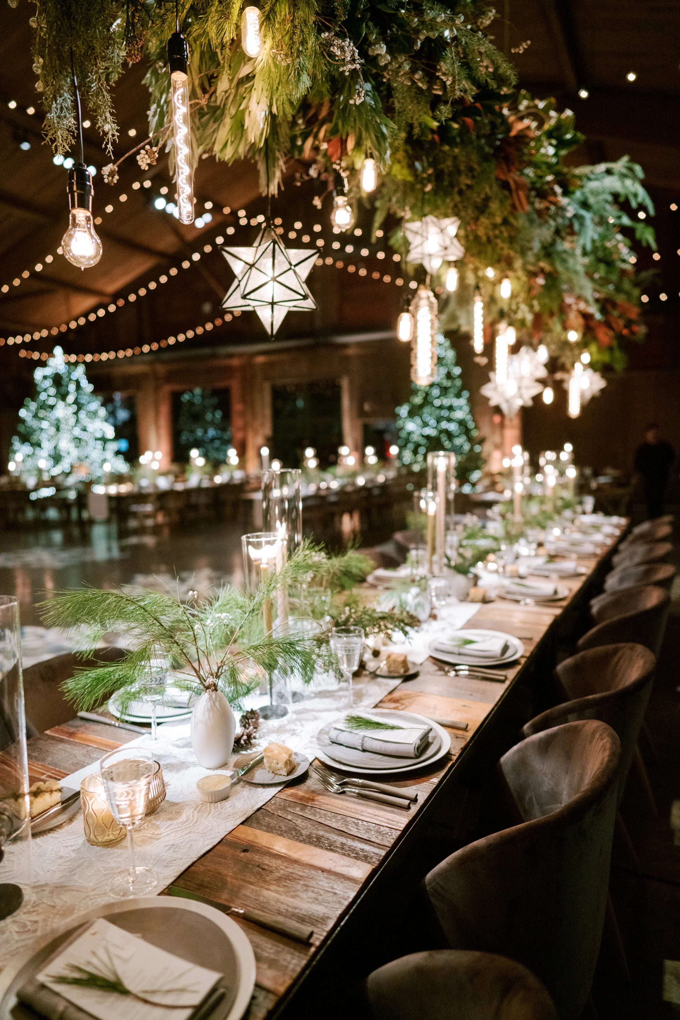 Festive holiday table decorated with greenery, candles, and place settings, with Christmas trees and string lights in the background.