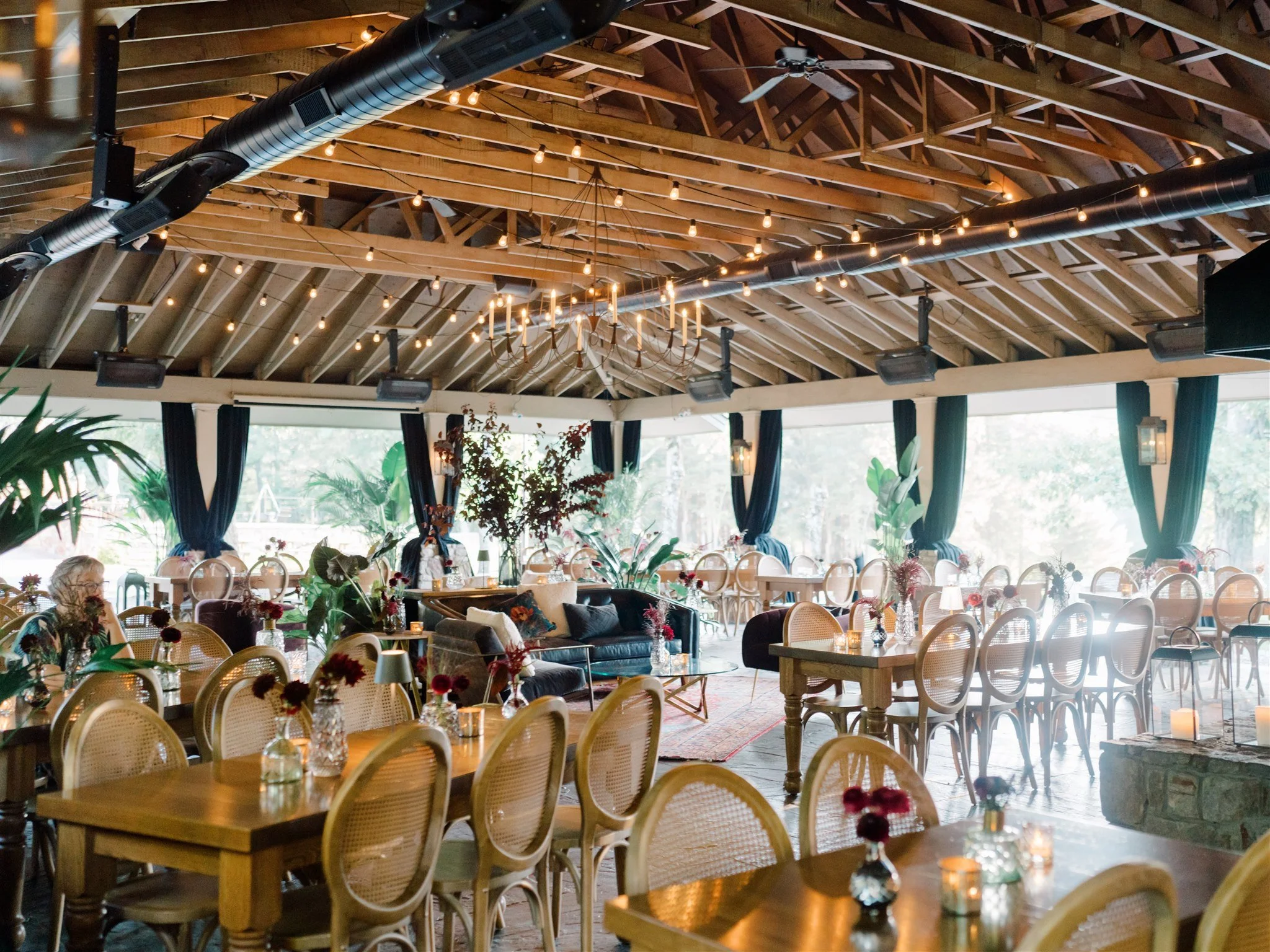 Elegant event space decorated with flowers, candles, and cozy furniture, featuring wooden tables, woven chairs, and large windows with dark curtains, illuminated by string lights and a chandelier.