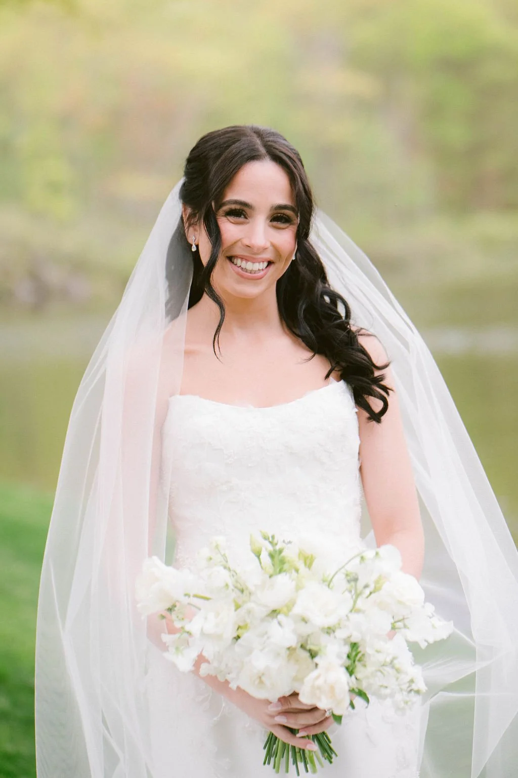 Smiling bride in white wedding dress holding a bouquet of white flowers outside with blurred greenery background.