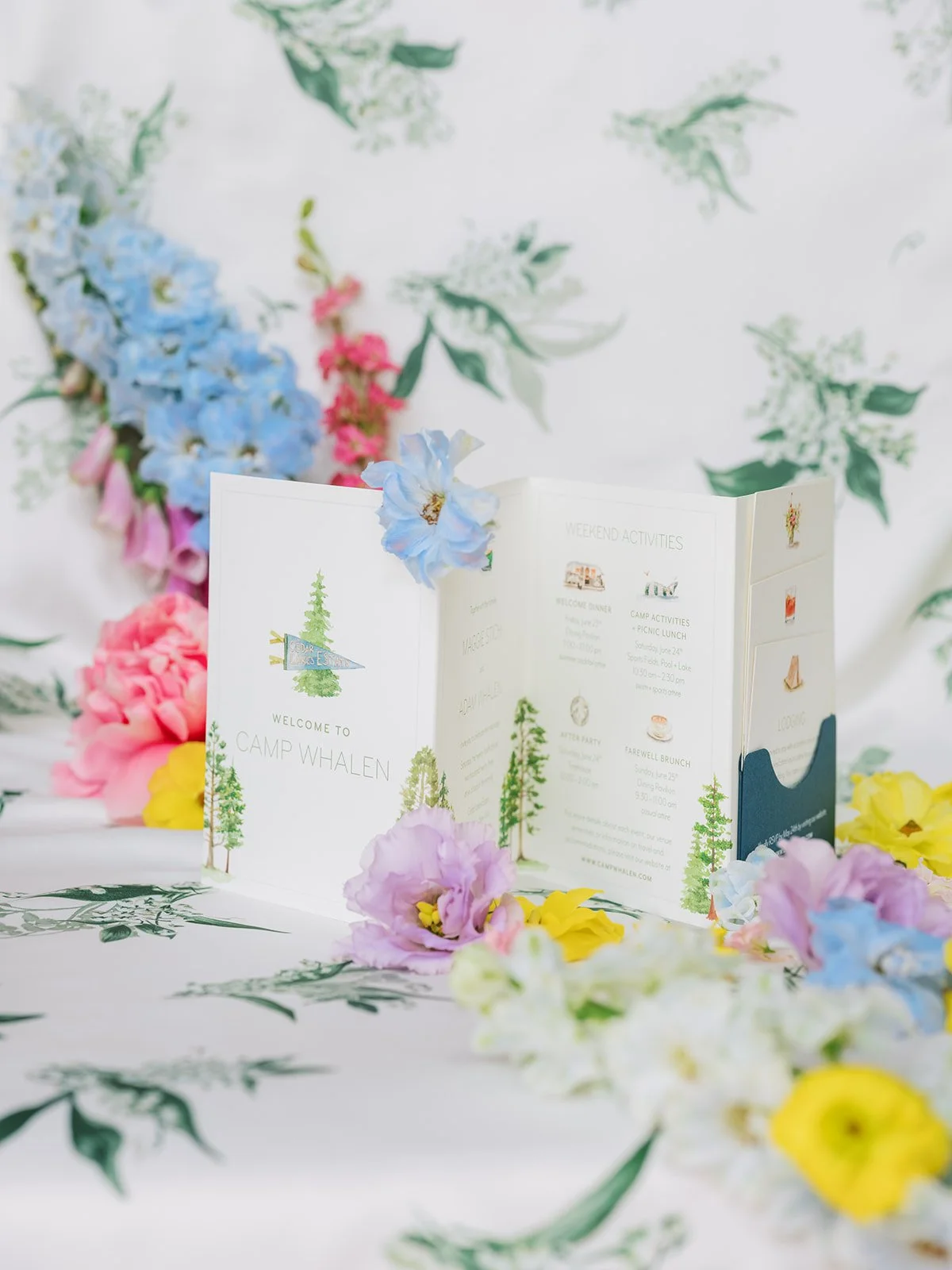 An open brochure on a white table surrounded by colorful flowers, with a floral backdrop, welcoming to Camp Whalen.