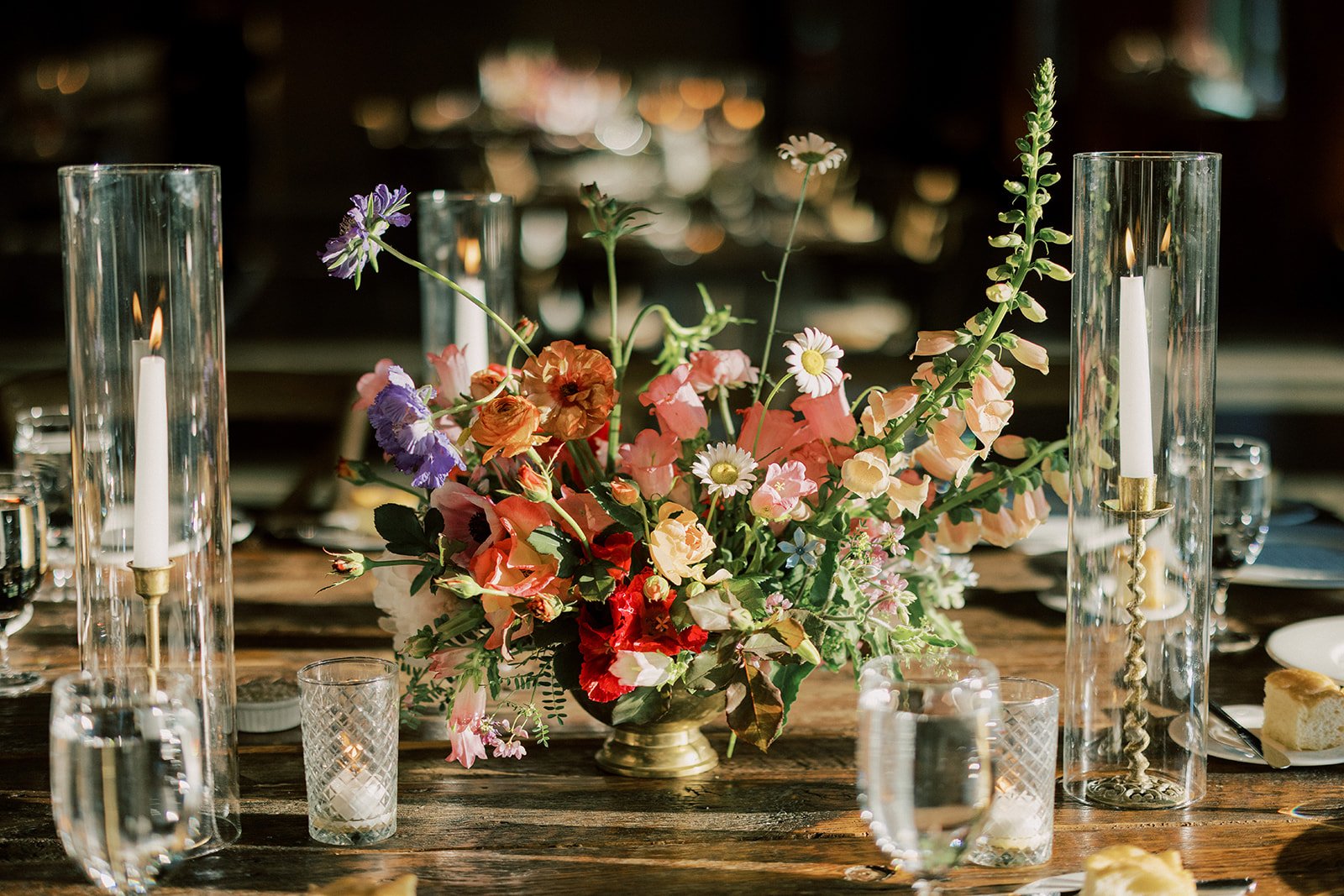 Elegant table centerpiece with a colorful flower arrangement in a gold vase, surrounded by tall glass candle holders with lit candles and smaller crystal glass holders, set on a rustic wooden table at a formal event.
