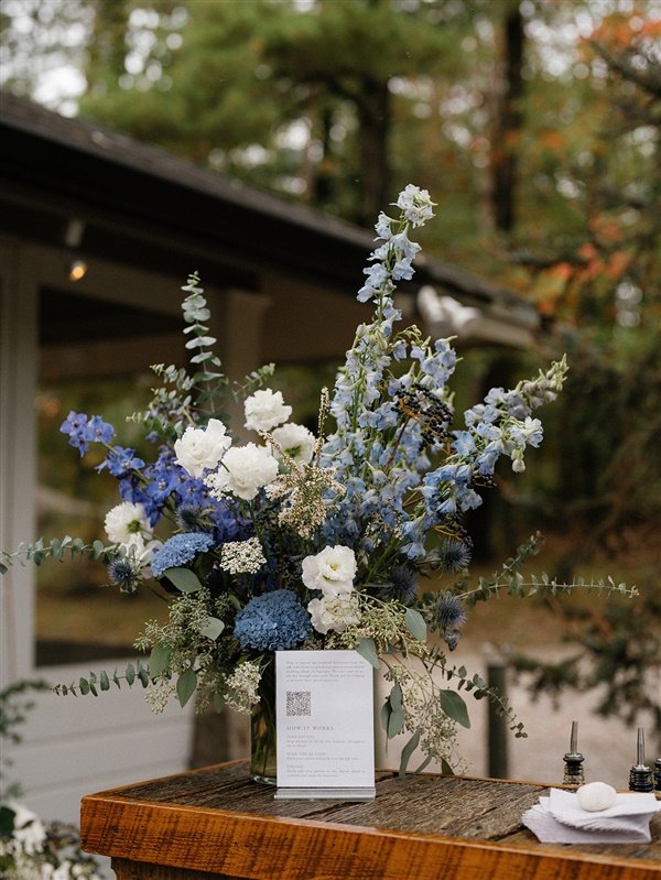 A floral arrangement with white, blue, and purple flowers and green foliage in a glass vase on a wooden surface outdoors, with trees and a building in the background.
