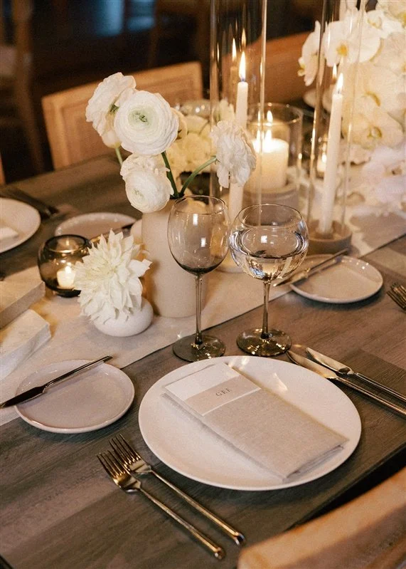 Elegant dining table setup with white flowers in vases, candles, wine glasses, and neatly folded napkins on white plates.