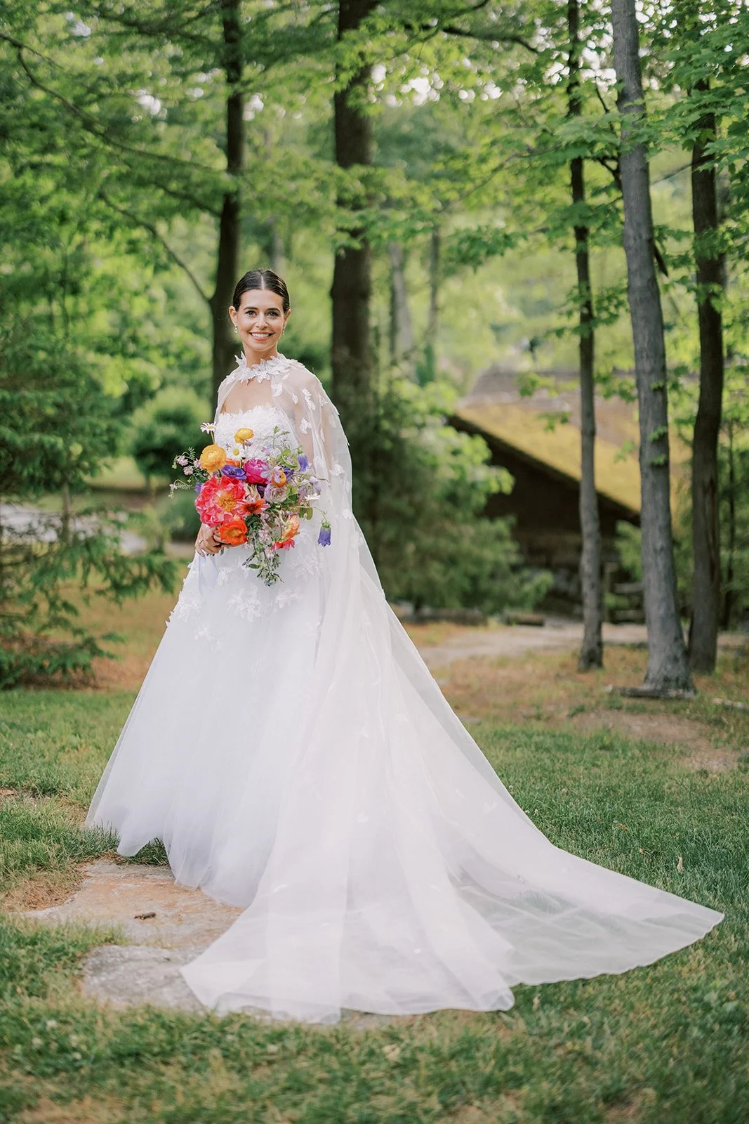 A bride in a white wedding gown holding a colorful bouquet of flowers, standing outdoors in a lush green forest.