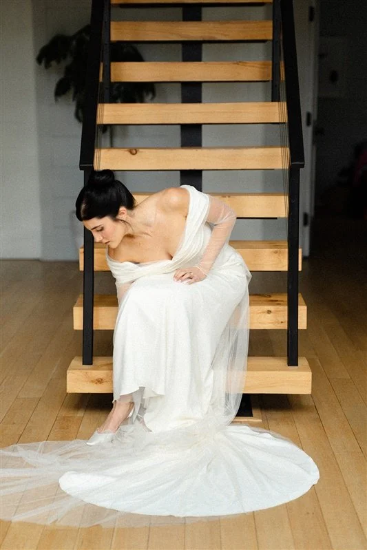 A woman in a white wedding dress sitting on stairs, looking down as she adjusts her shoe.