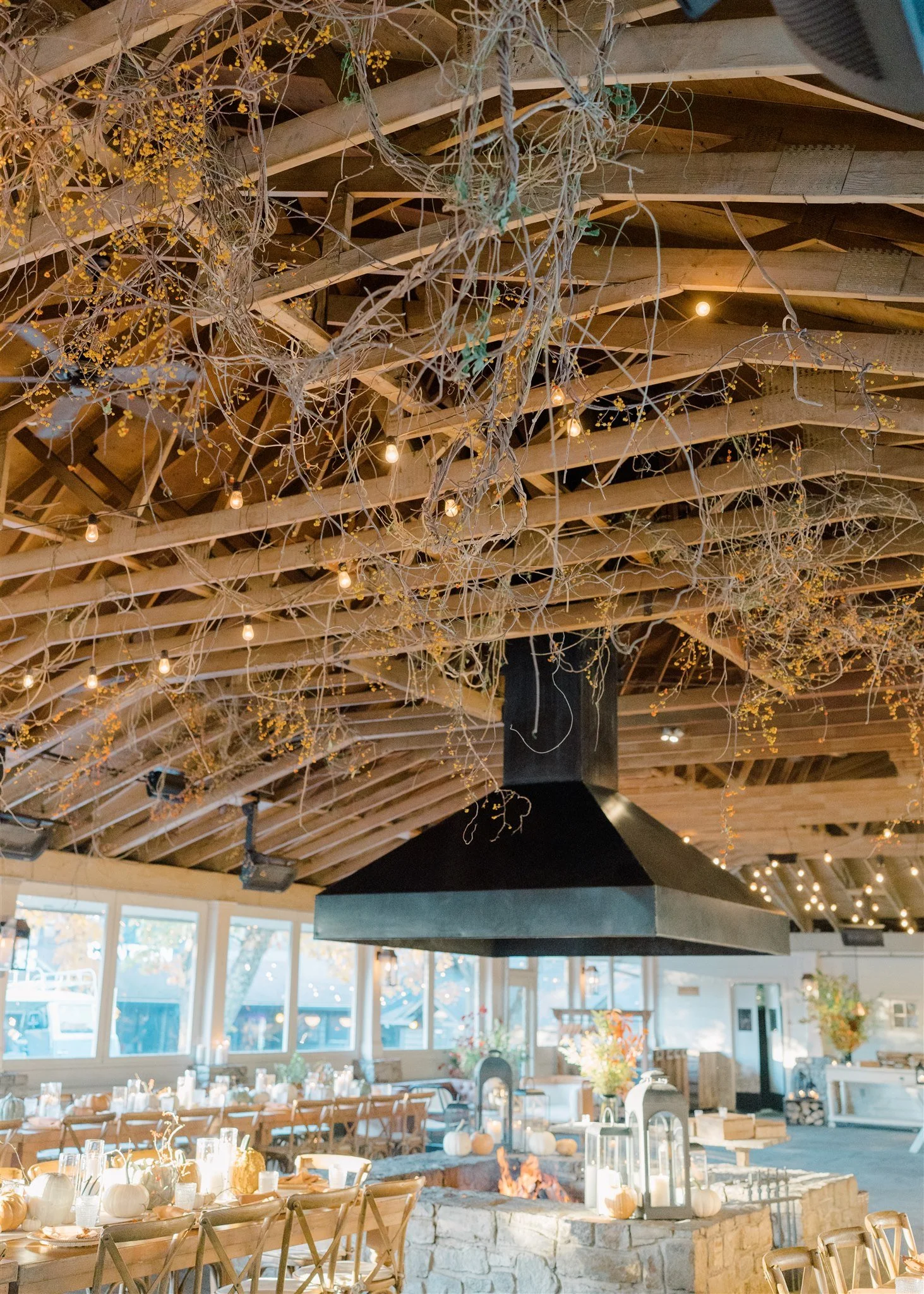 Interior of a decorated event hall featuring a wooden ceiling with hanging dried branches and string lights. A circular stone fire pit decorated with pumpkins and lanterns is in the center, surrounded by tables and chairs, with natural light coming t