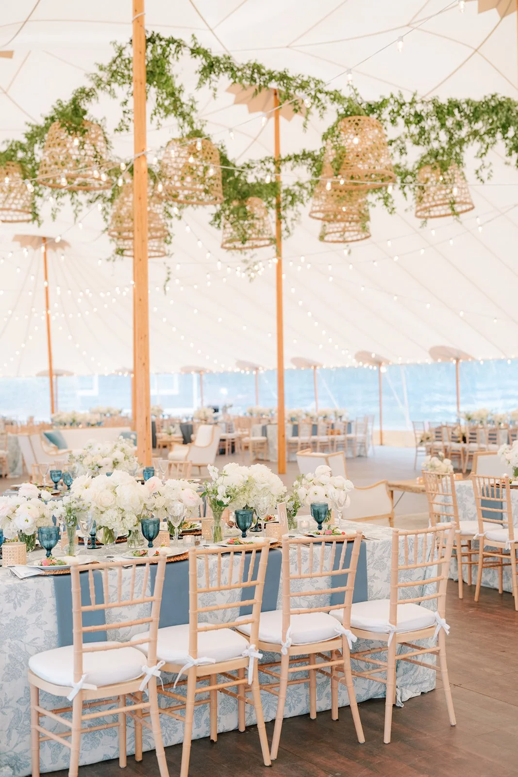 A decorated event space with tables set for a celebration, featuring floral centerpieces, blue glassware, and wooden chairs. The ceiling is adorned with string lights, green drapes, and woven lanterns, with a view of water and sky in the background.