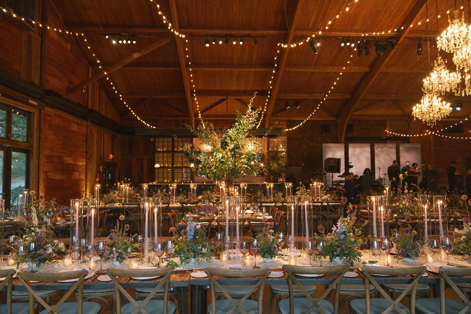 A decorated banquet hall with long tables, floral centerpieces, lit candles, and string lights on the wooden ceiling, with a band performing in the background.