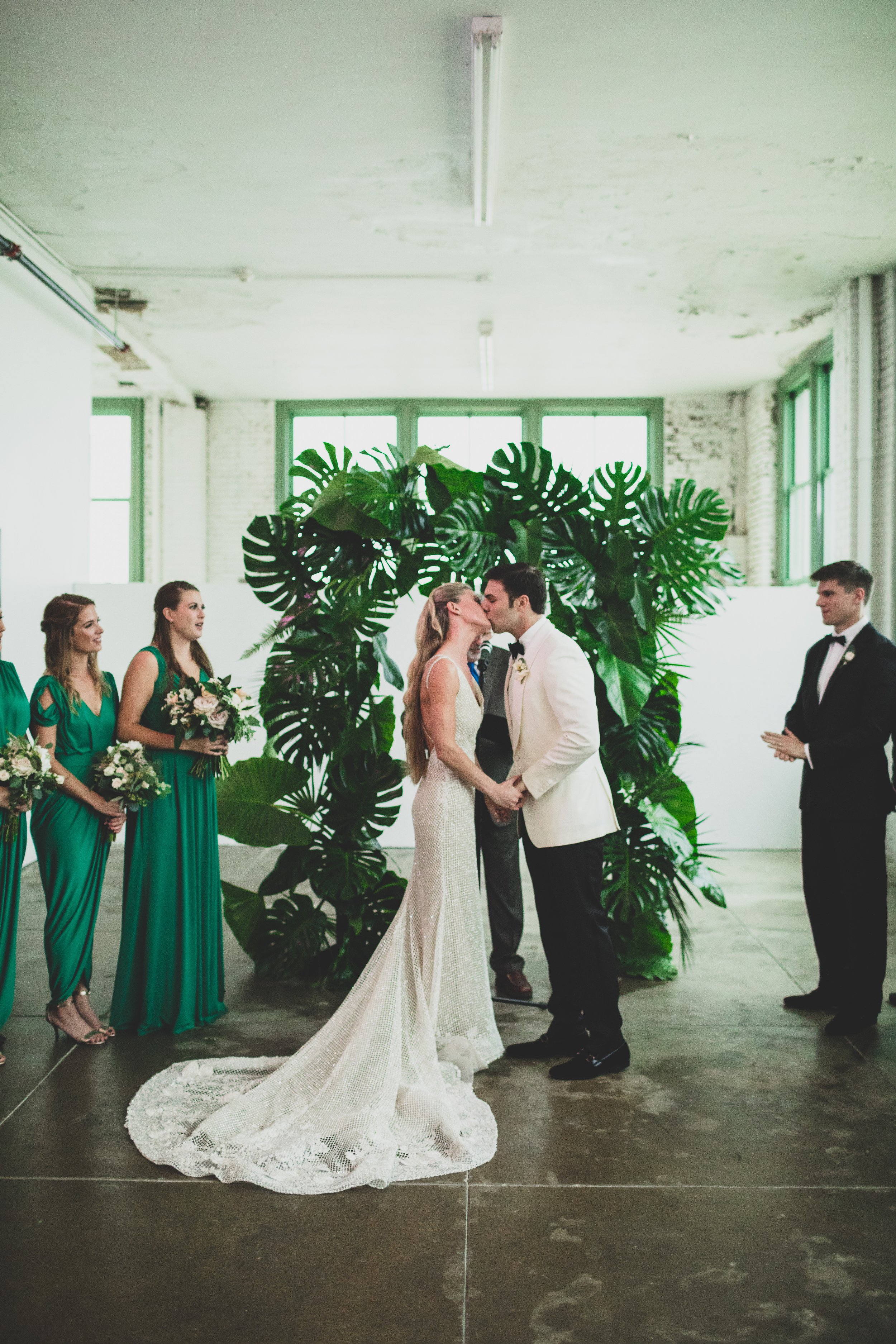 A wedding ceremony inside with a couple kissing in front of a green leafy backdrop, surrounded by bridesmaids in teal dresses and groomsmen in black tuxedos.