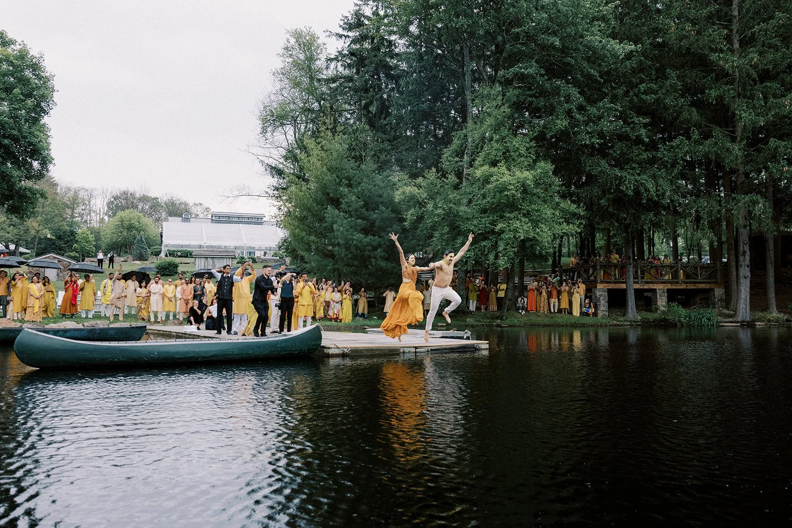 A wedding party on a dock by a lake with a large group of guests, and a bride and groom jumping into the water as part of the celebration, surrounded by trees and outdoor scenery.