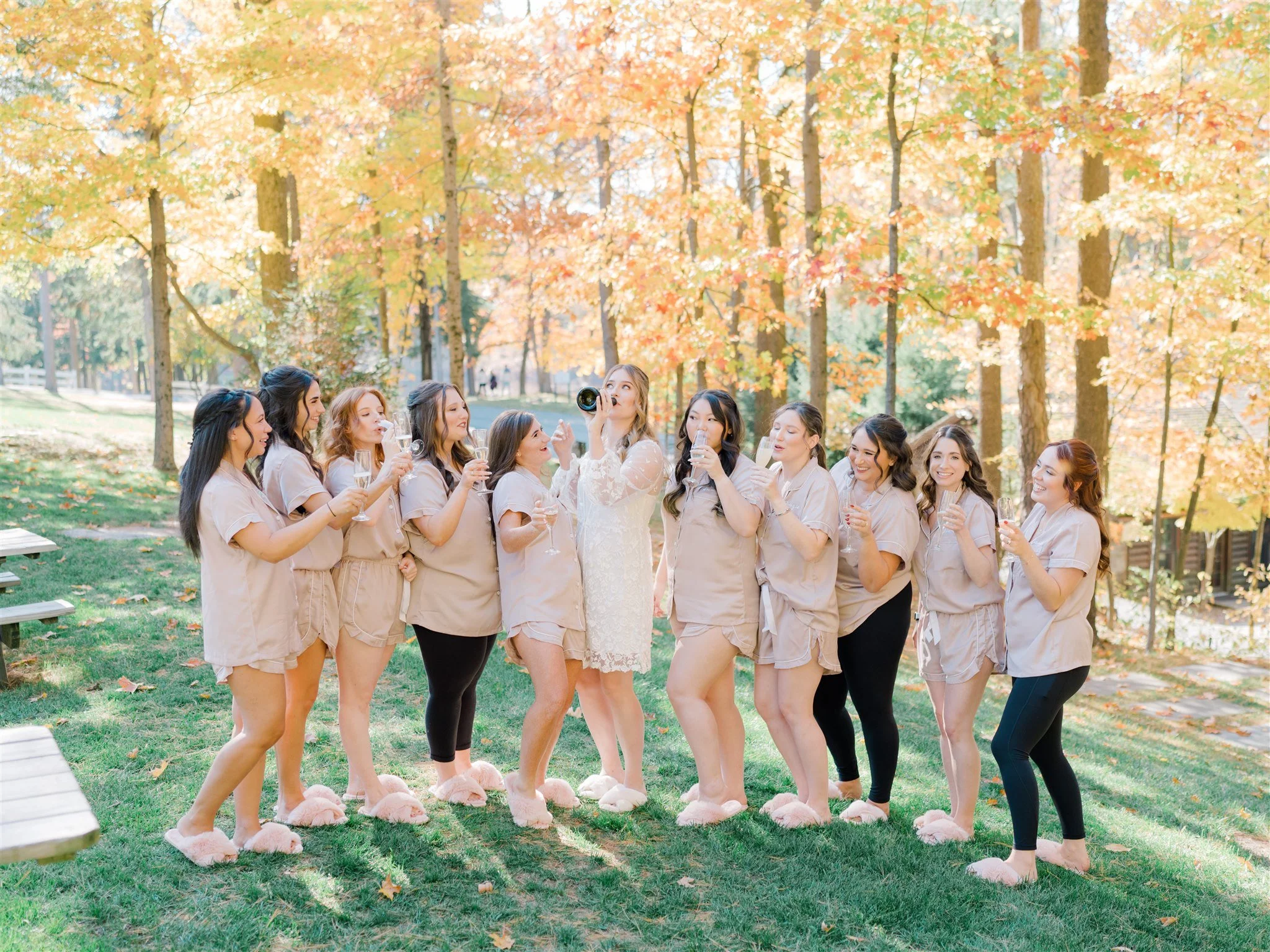 A bridal party of twelve women dressed in beige pajamas and fluffy pink slippers celebrating outdoors in a park with fall foliage, holding glasses of champagne, with some women taking photos and enjoying the moment.