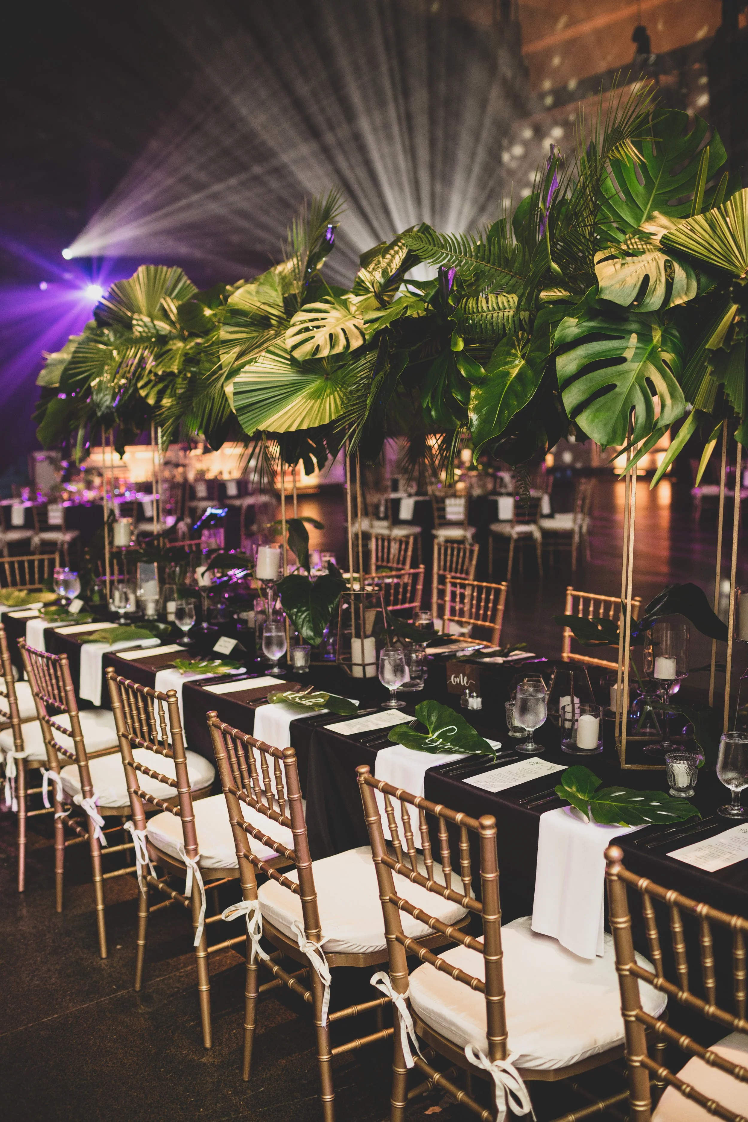 Elegant event space decorated with lush green tropical plants, set with black and white table settings, gold chairs, and purple lighting.