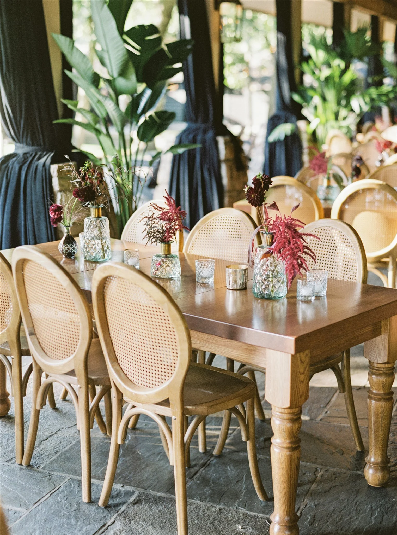 A wooden dining table decorated with vases of pink and red flowers and small glass candle holders, in a bright room with large leafy plants and blue curtains.