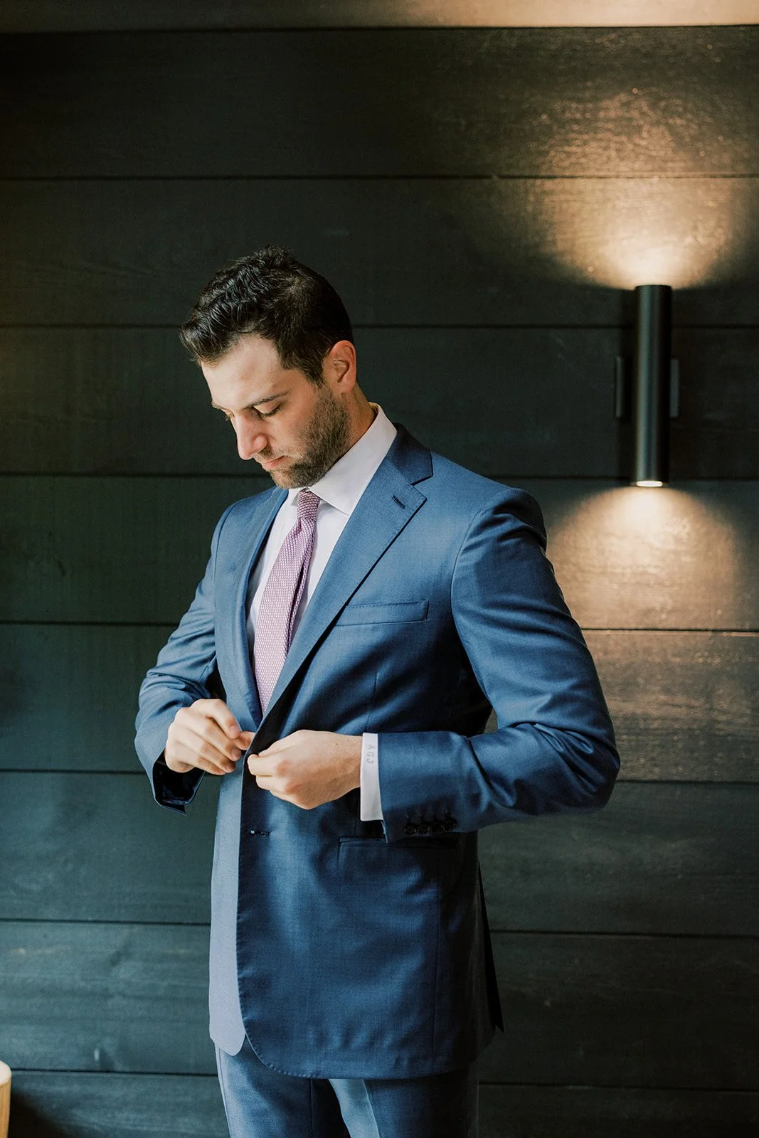 Man in a blue suit buttoning his jacket, standing against a dark wood-paneled wall with modern wall sconces.