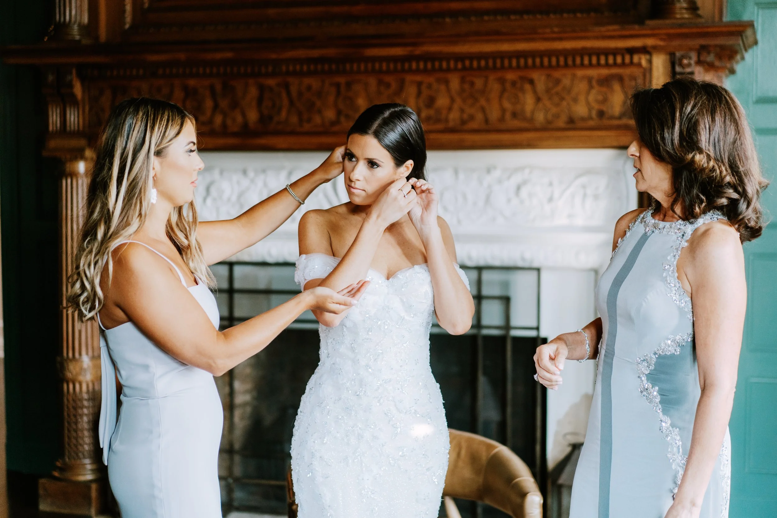 A woman in a wedding dress is being helped by two other women as she prepares for her wedding in an elegant room with a fireplace.