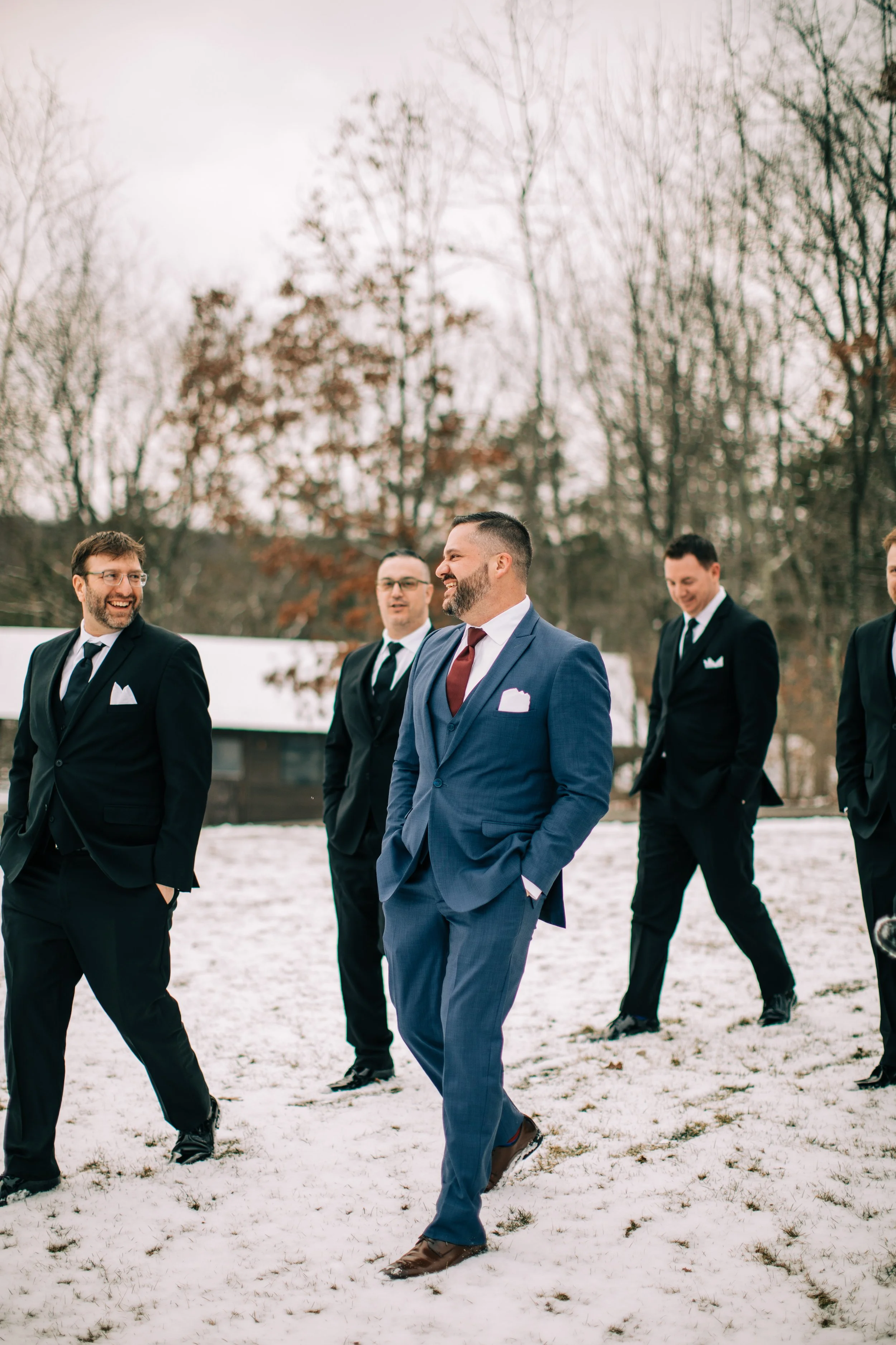 Group of men in suits walking outdoors on snow-covered ground, smiling and talking, with leafless trees and a barn in the background.