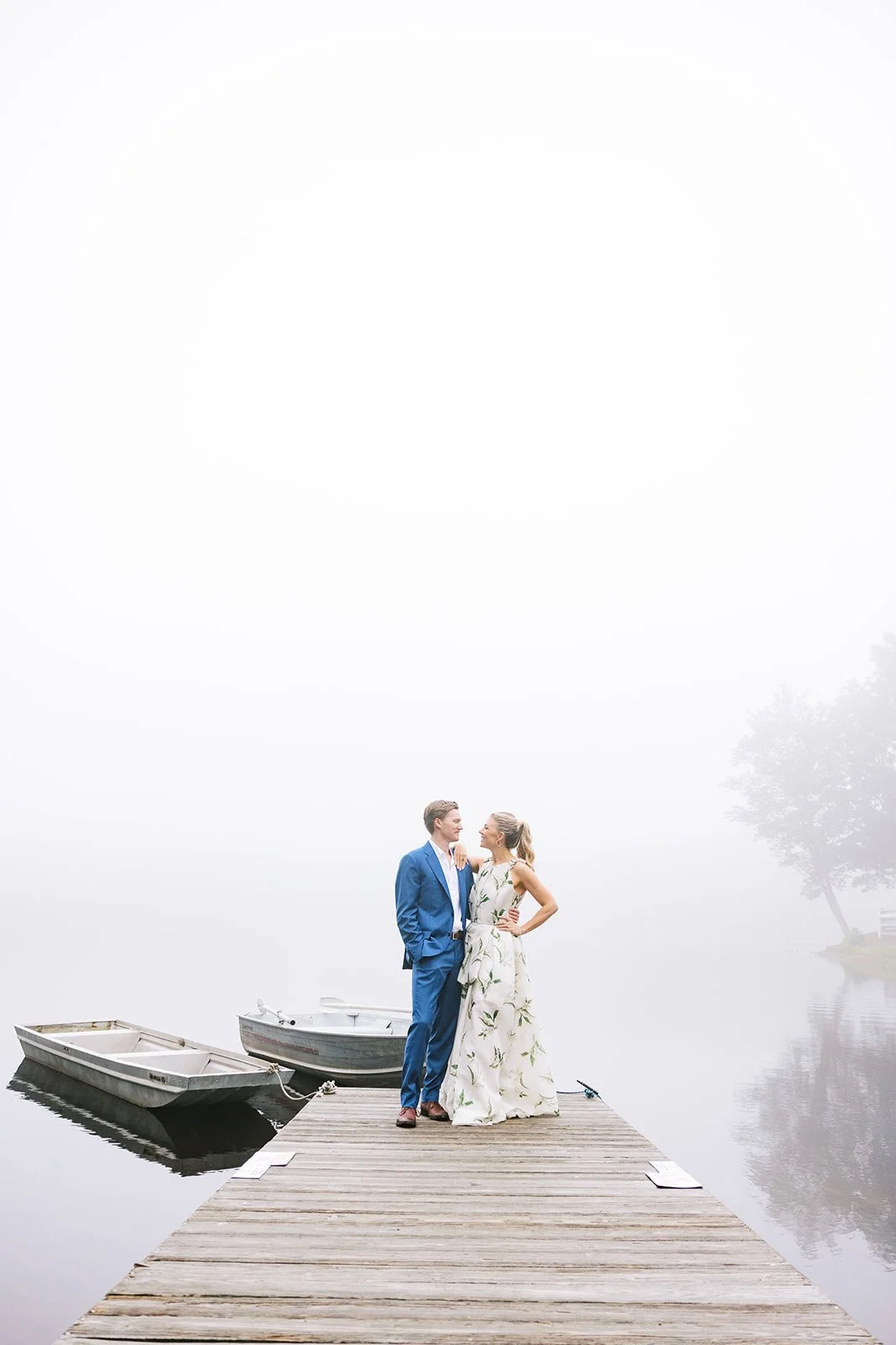 A man and woman in formal attire standing on a wooden dock by a foggy lake, with boats and trees visible in the background.