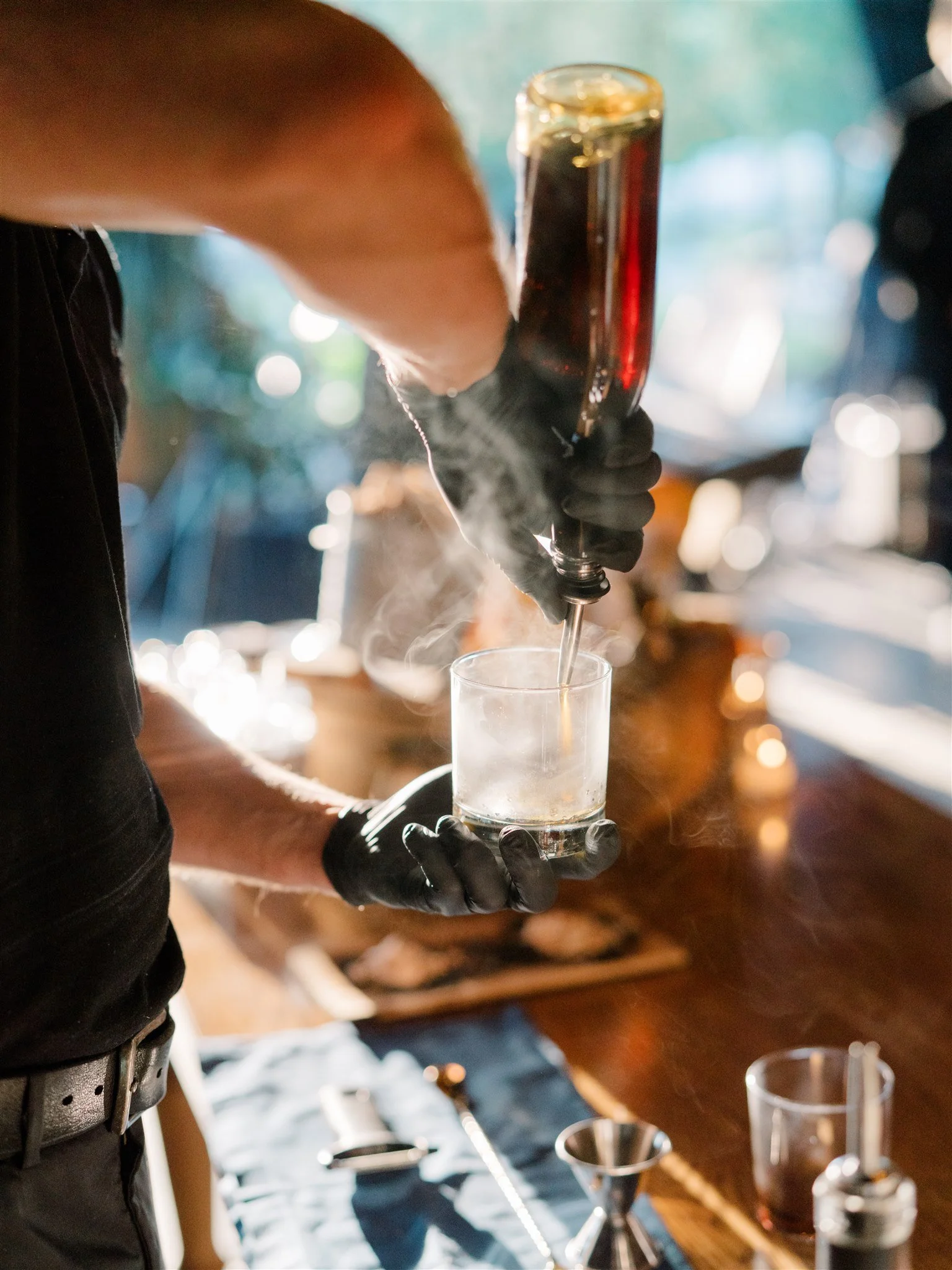 Bartender pouring a dark beverage into a glass with dry ice, creating fog, at a bar with bar tools on the counter.