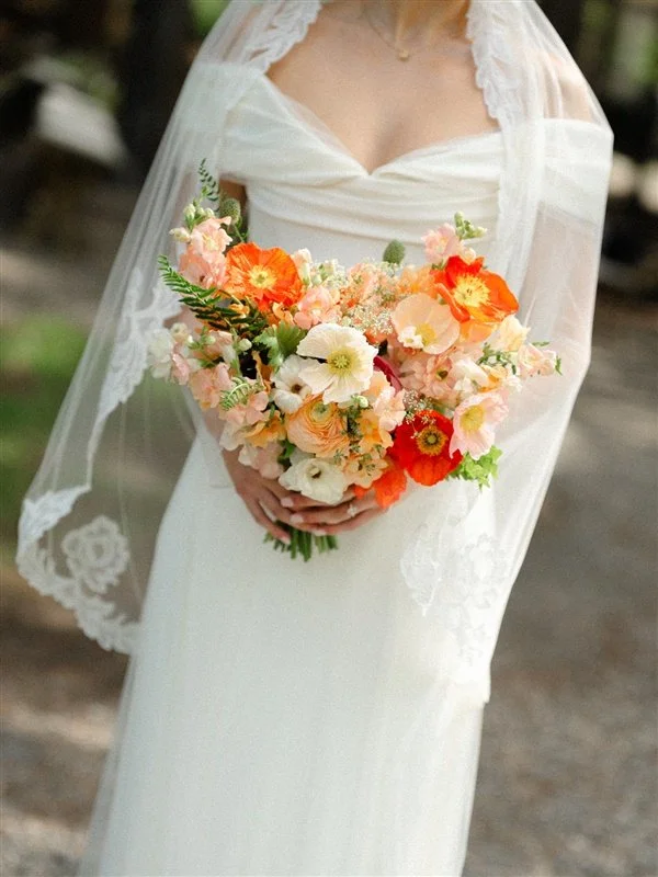 A bride in a white wedding dress holding a large bouquet of mixed flowers, including poppies and anemones, with a lace veil and a blurred outdoor background.
