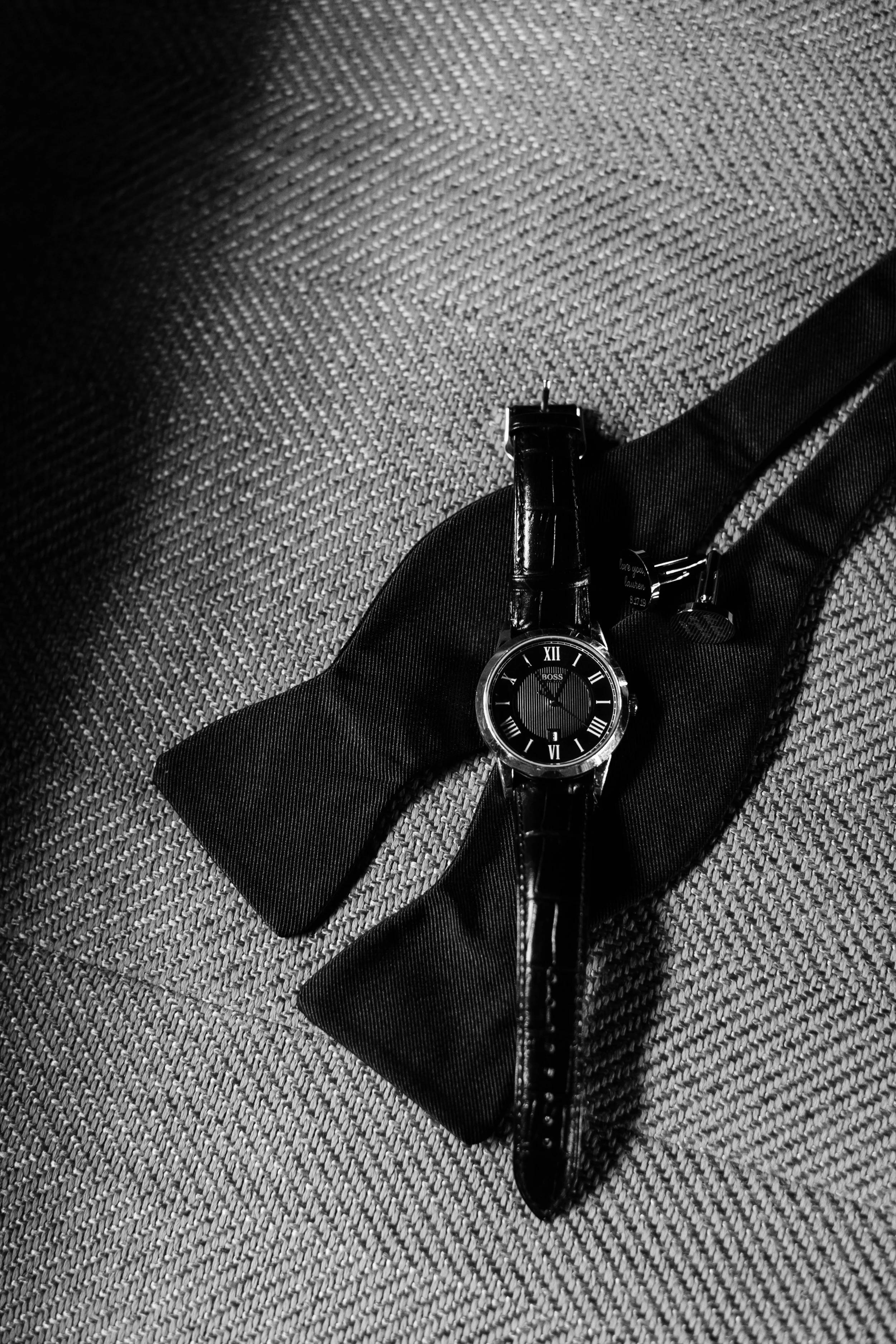 Black wristwatch with Roman numerals on a textured surface, resting on a black bow tie.