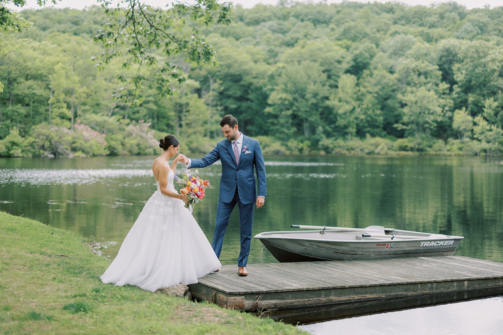 A bride and groom standing on a wooden dock by a lake, with the bride holding a bouquet of flowers and the groom wearing a blue suit, near a boat on the water, surrounded by lush green trees.