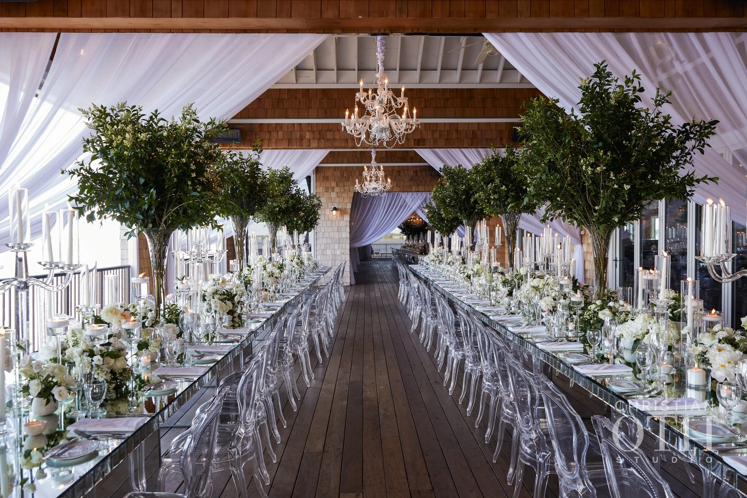 Elegant banquet hall decorated with white flowers, candleholders, and chandeliers, with a long wooden floor and draped ceiling.