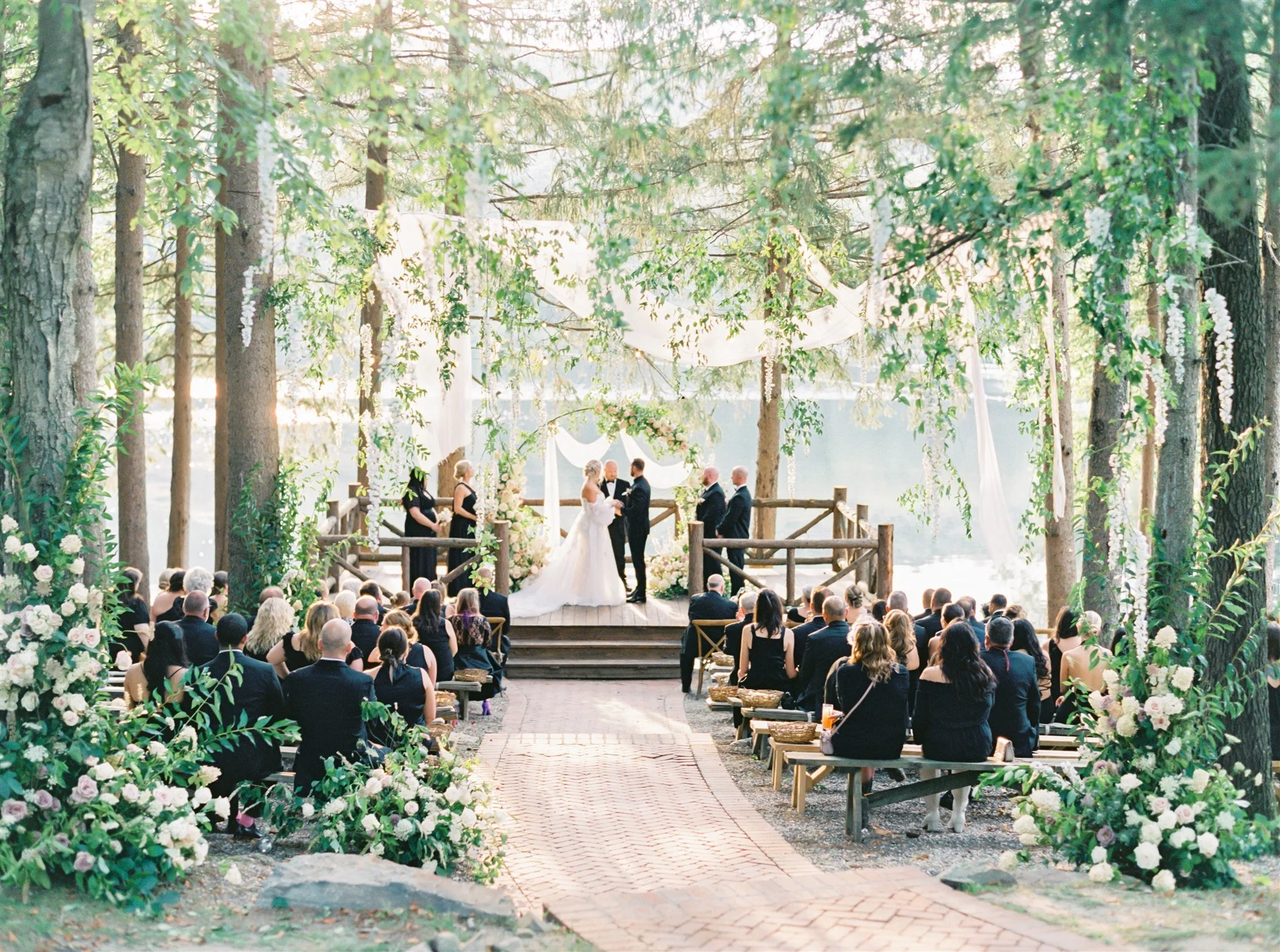 A wedding ceremony taking place outdoors in a wooded area by a lake. The bride and groom are exchanging vows on a wooden platform decorated with flowers and white drapes, with guests seated in front.