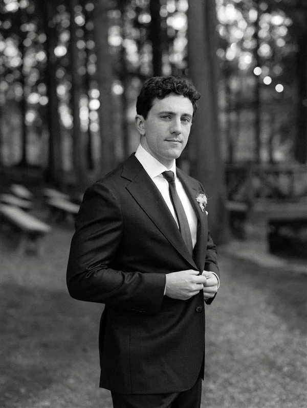 A young man in a dark suit, white shirt, and tie standing outdoors in a wooded area, adjusting his jacket.