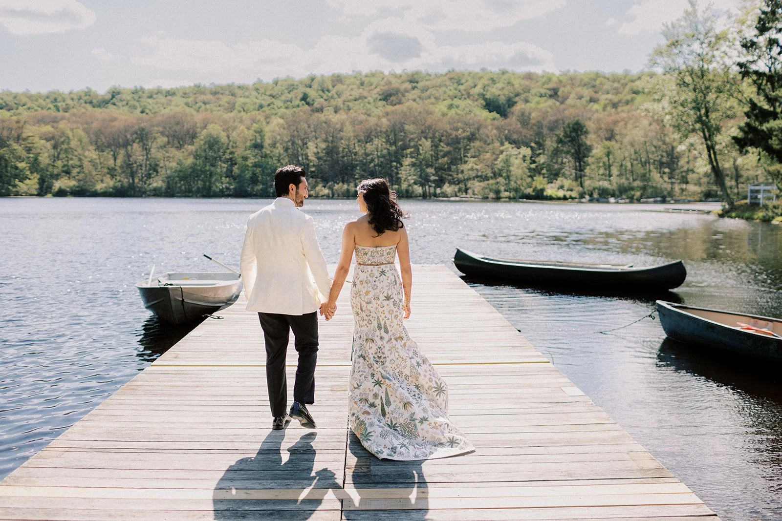 A couple holding hands walking on a wooden dock by a lake with three boats and a lush green forest in the background.